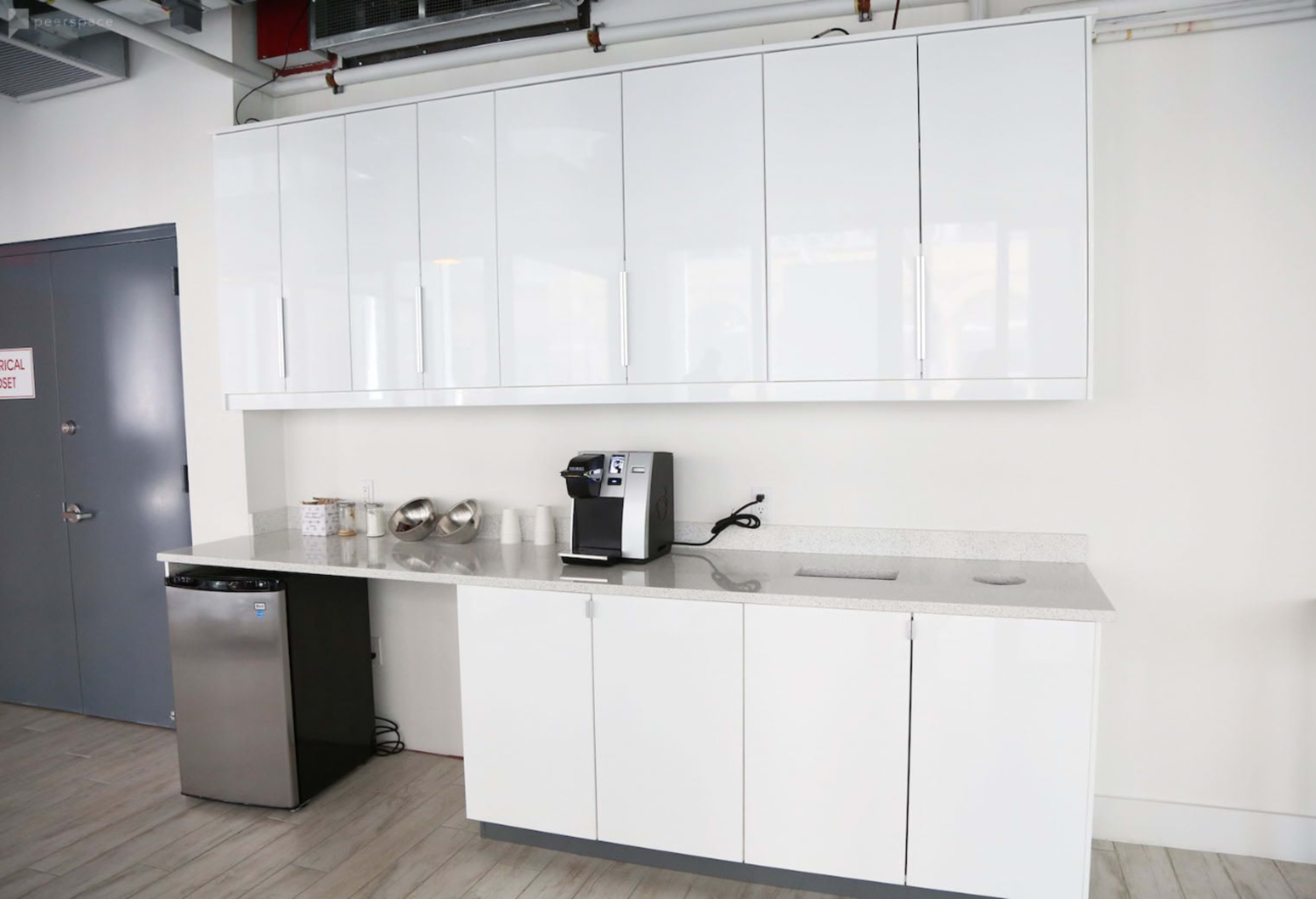The image shows a modern kitchen area with white cabinets, a countertop, a coffee machine, and a small refrigerator.
