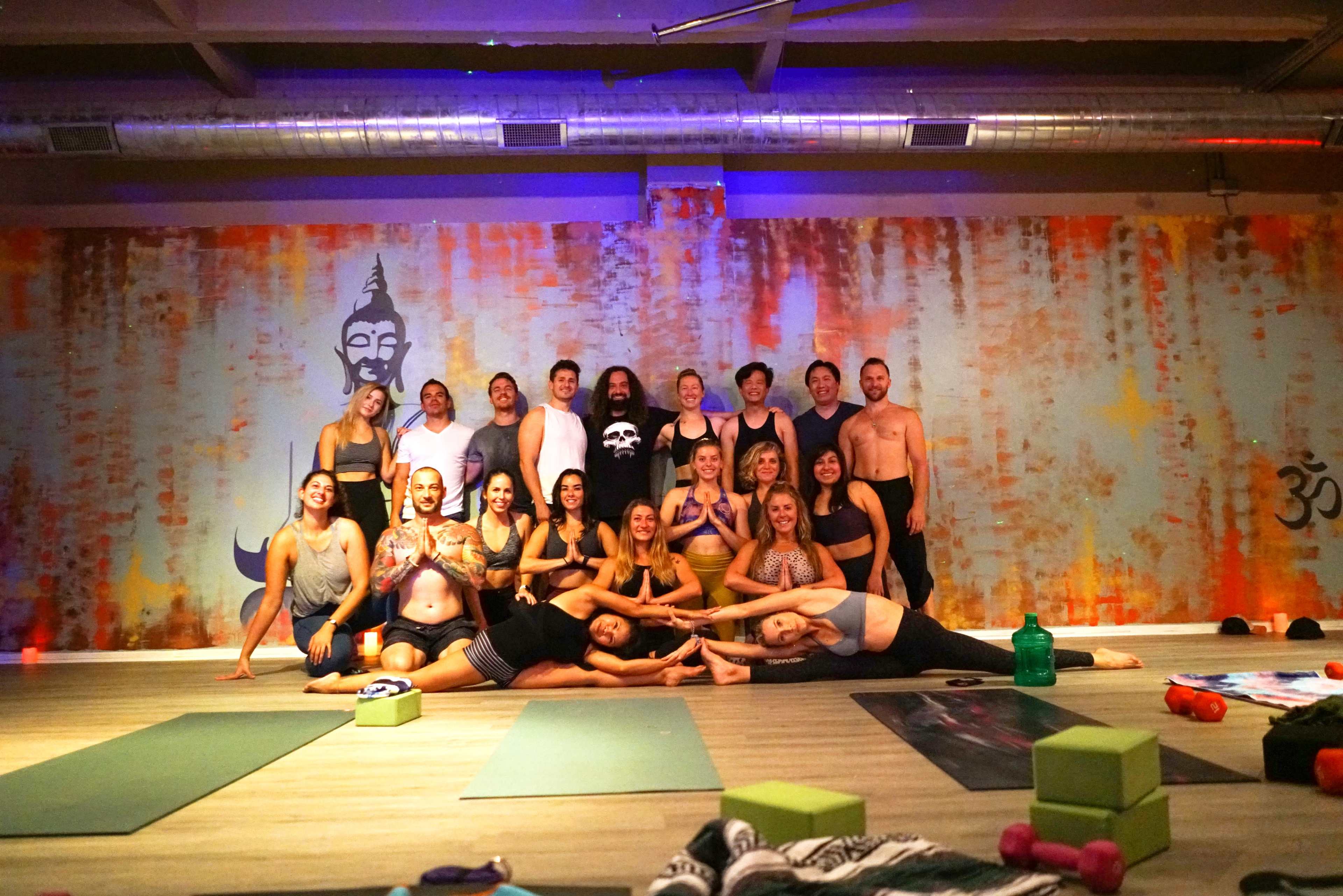 A group of twenty people poses together in a yoga studio with colorful wall art and yoga mats on the floor.