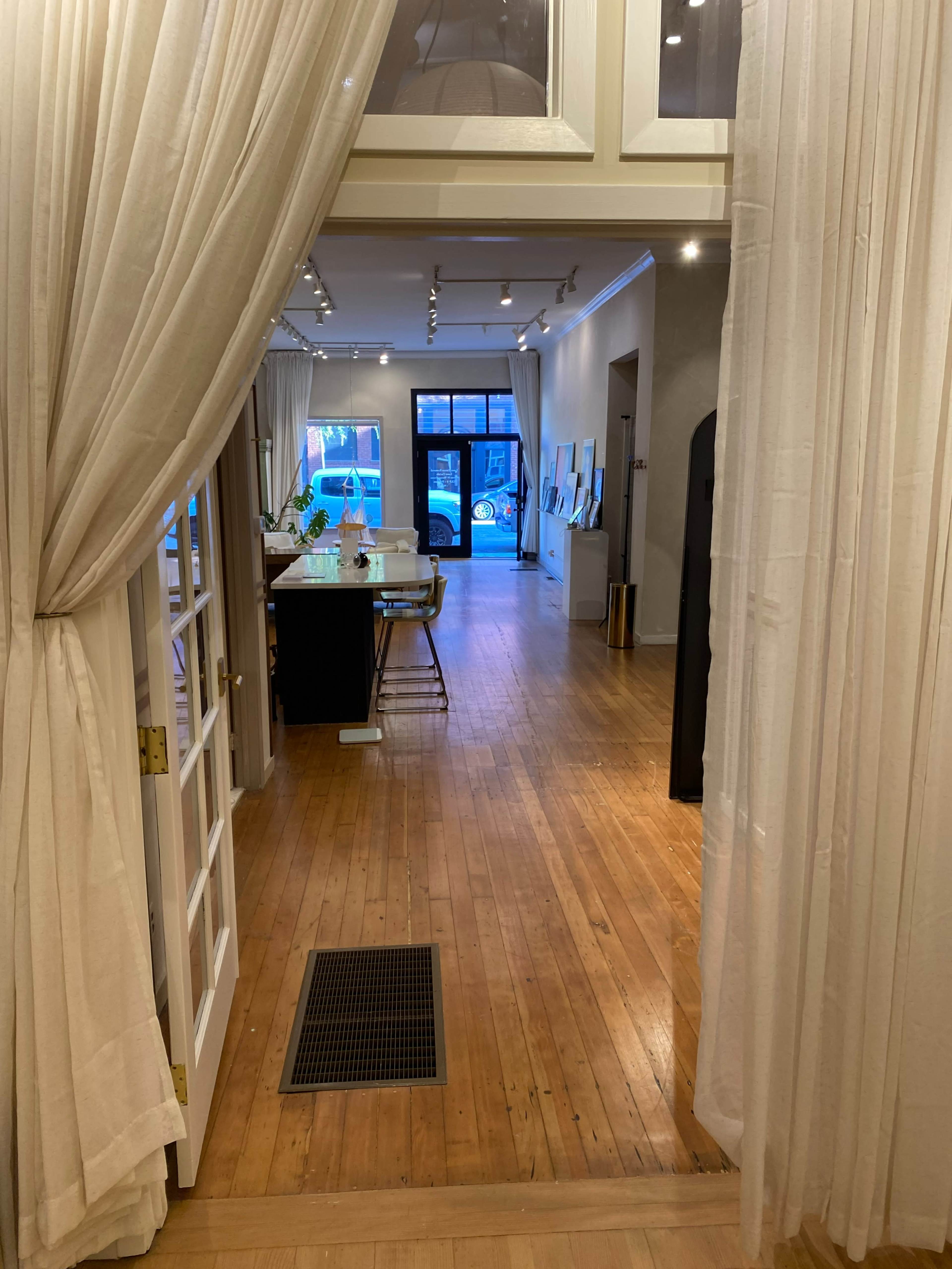 The image shows a view of a spacious, well-lit interior leading to an open area with a dining table and various artworks displayed on the walls.
