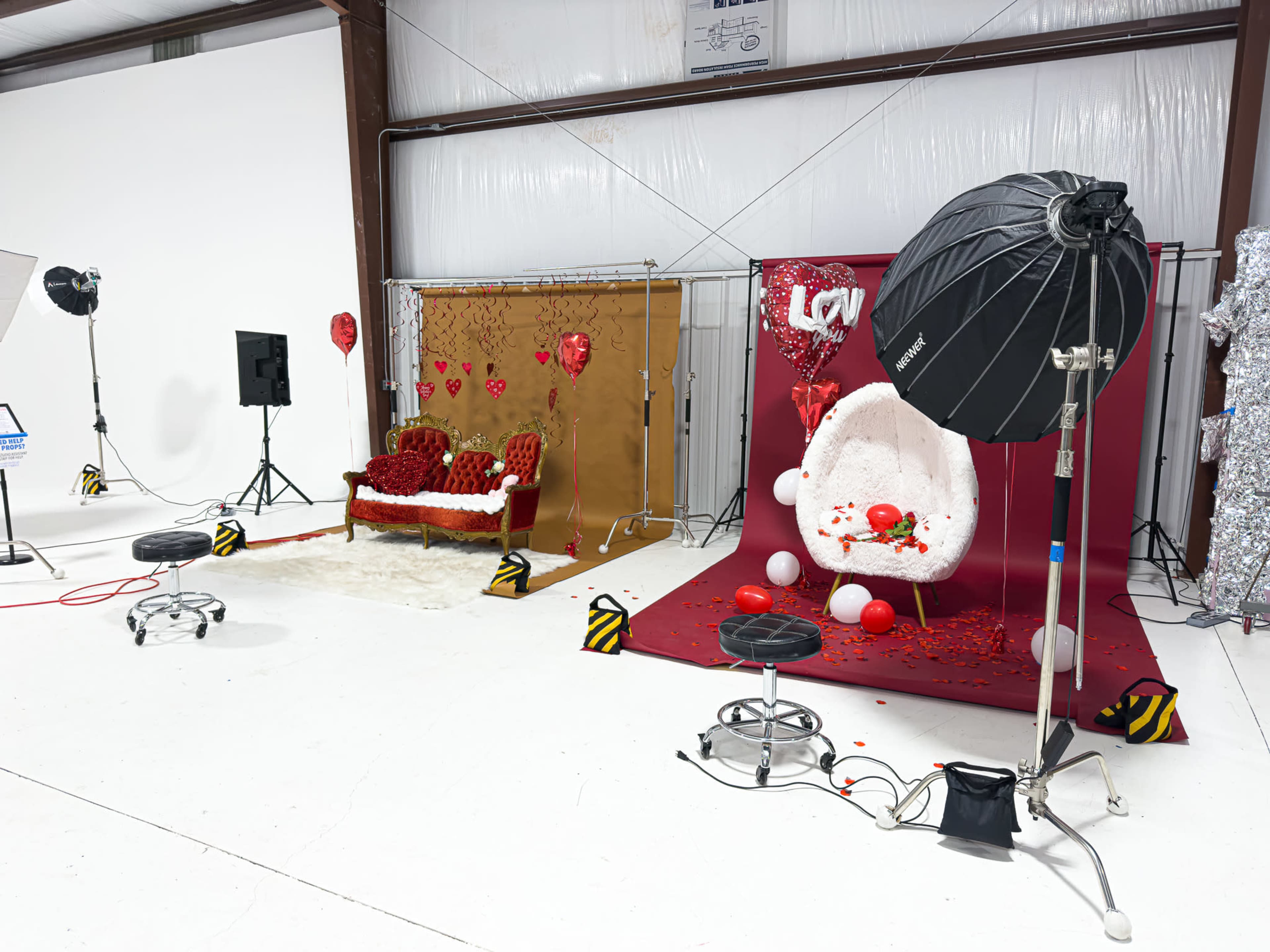 The image shows a photography studio setup with a red and white theme, featuring a sofa adorned with heart decorations, a white chair, and props like balloons and scattered rose petals.