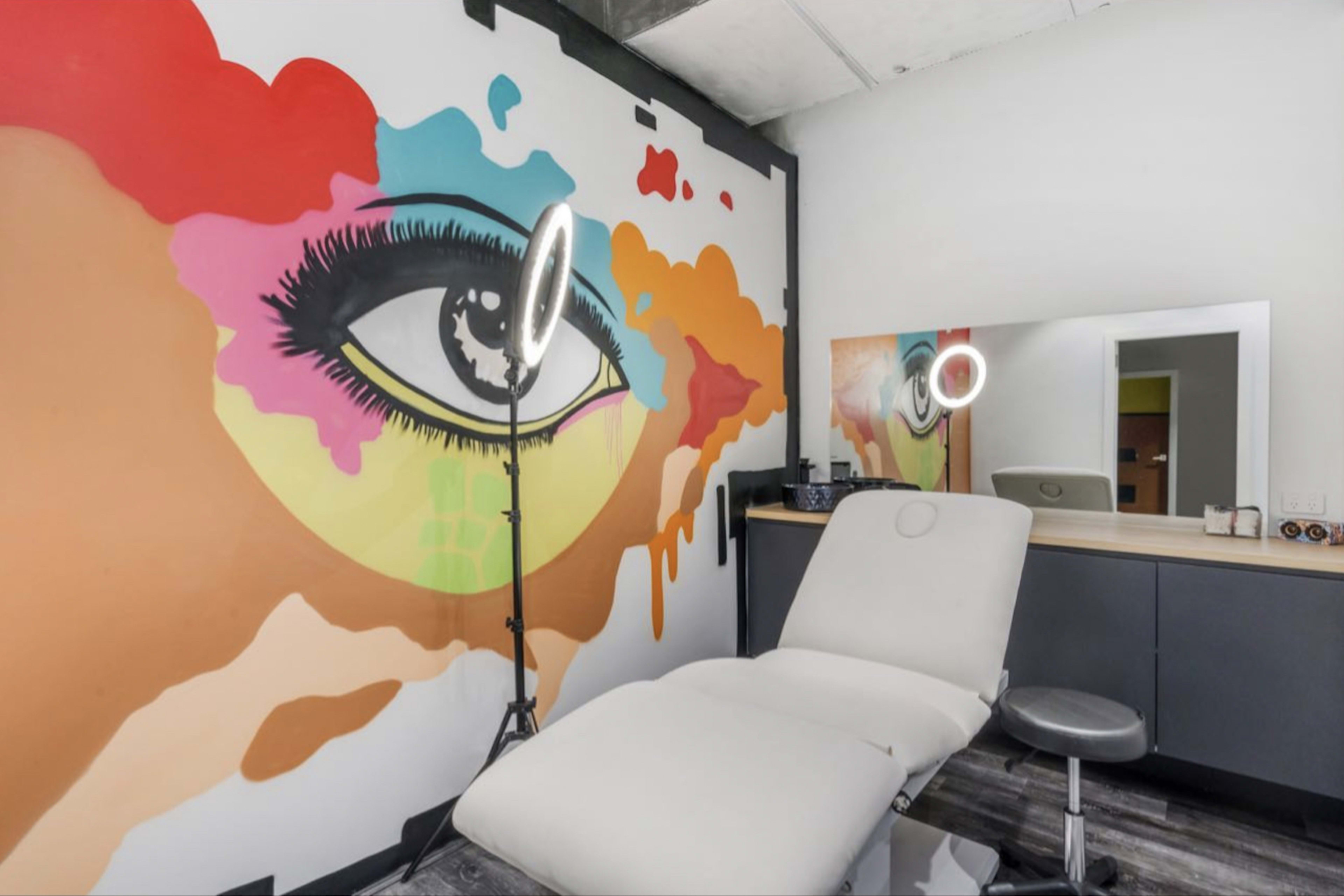 A beauty treatment room featuring a large colorful mural of an eye on the wall, a treatment chair, and a mirror with a ring light.