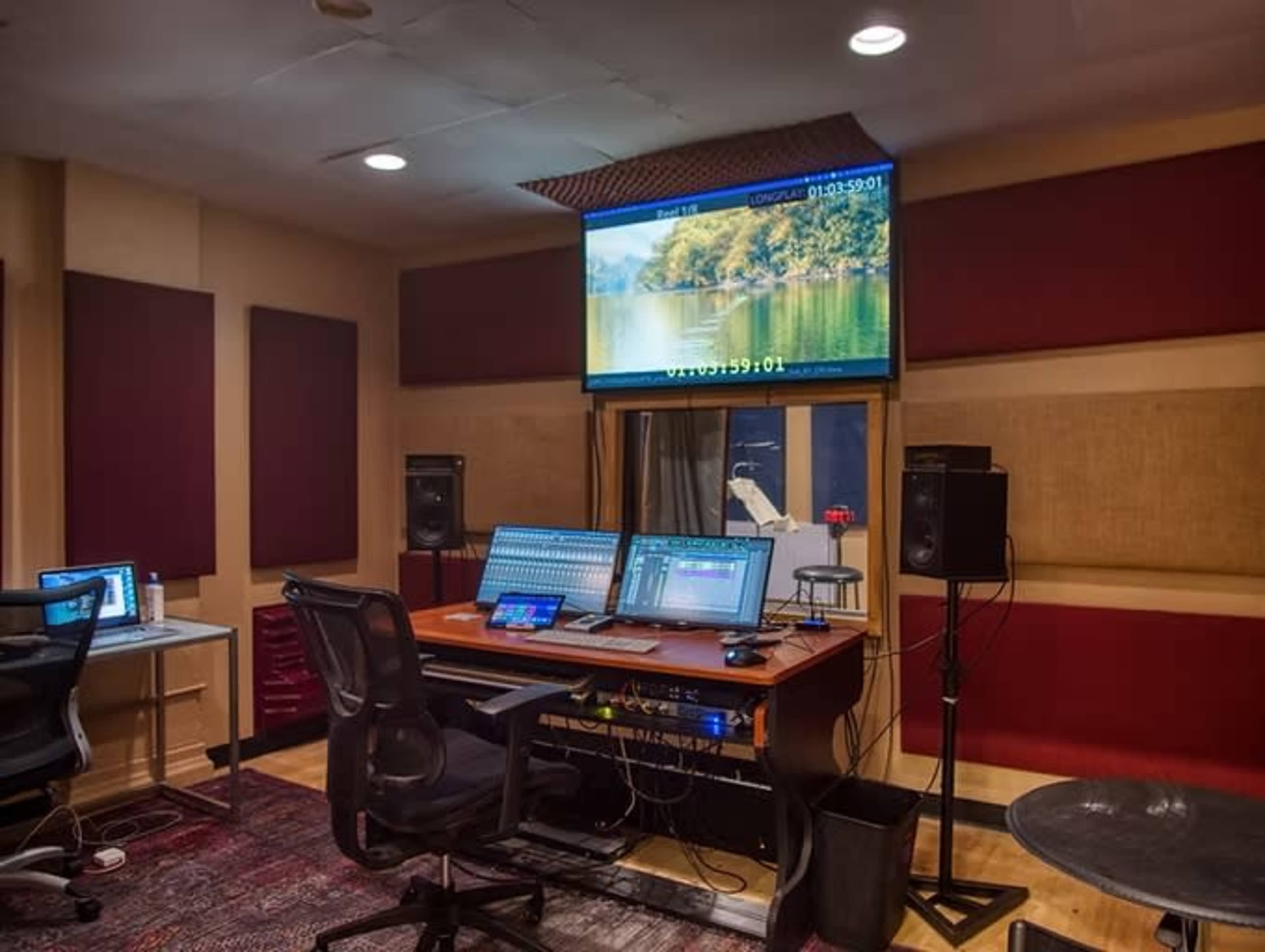 The image shows a sound recording studio featuring a large mixing desk, computer monitors, speakers, and soundproof wall panels.