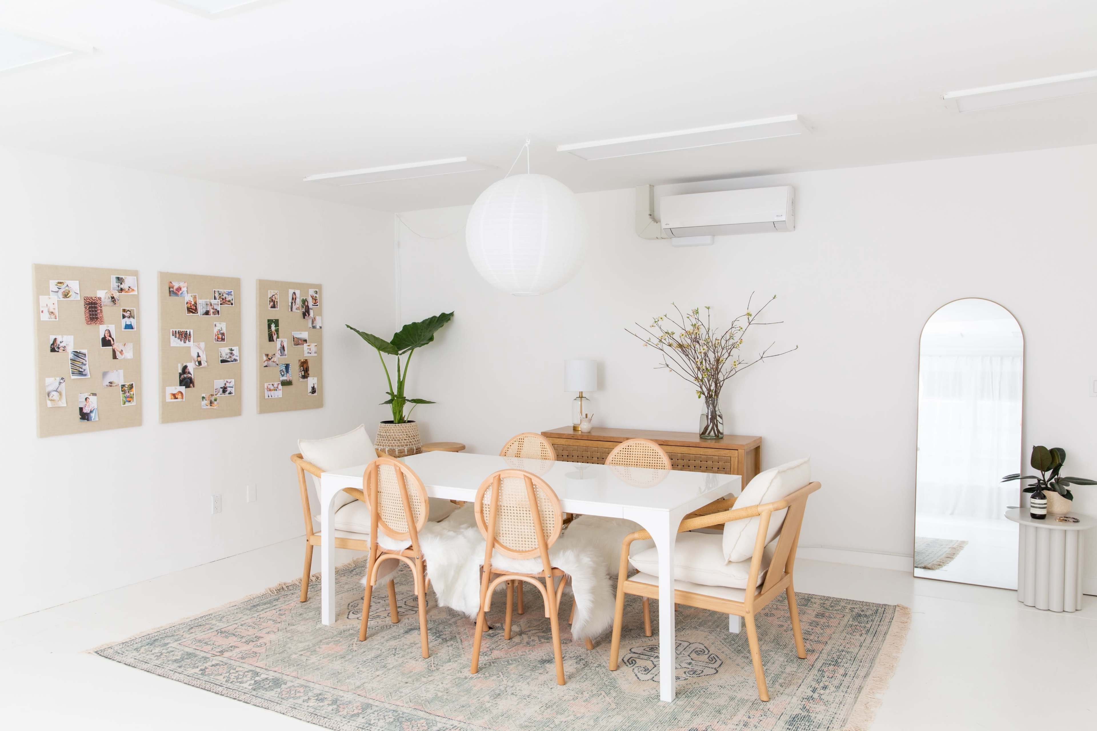 A bright, modern dining room features a white table surrounded by wooden chairs, a gallery wall of photos, and a large mirror.