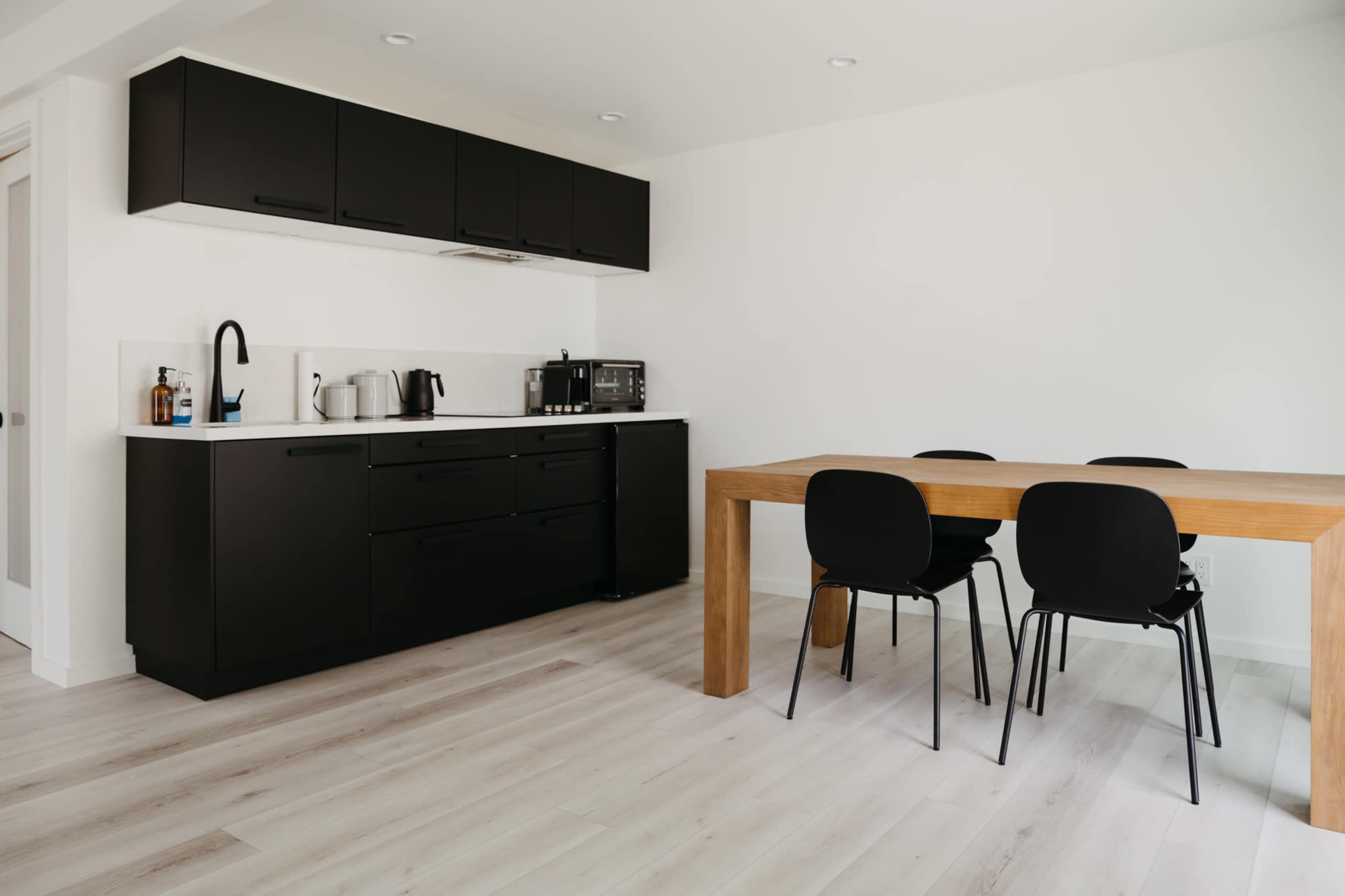 A modern kitchen with black cabinets, a white countertop, and a wooden dining table surrounded by four black chairs.