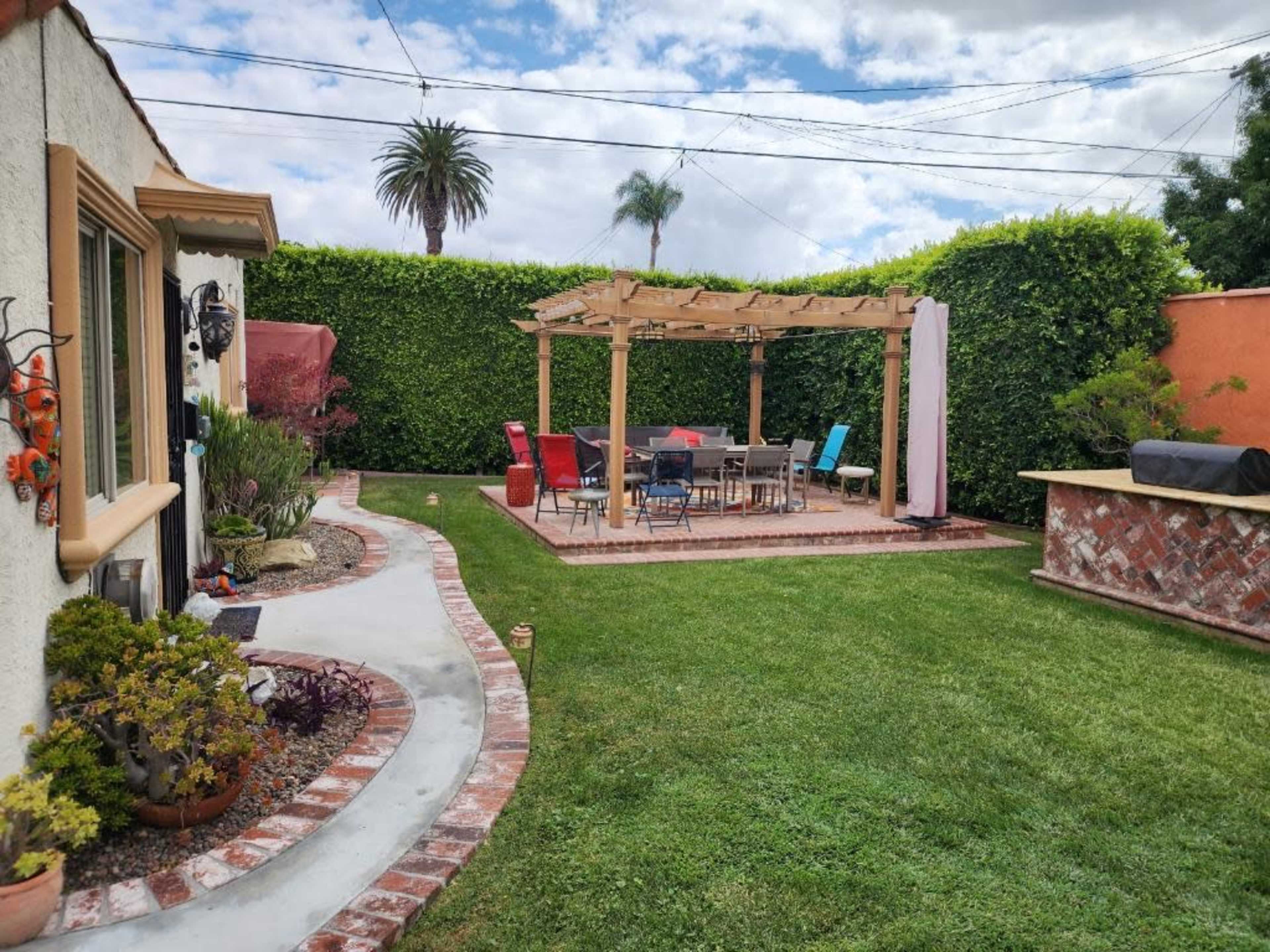 The image shows a backyard with a wooden pergola over a dining area surrounded by well-manicured grass and hedges.