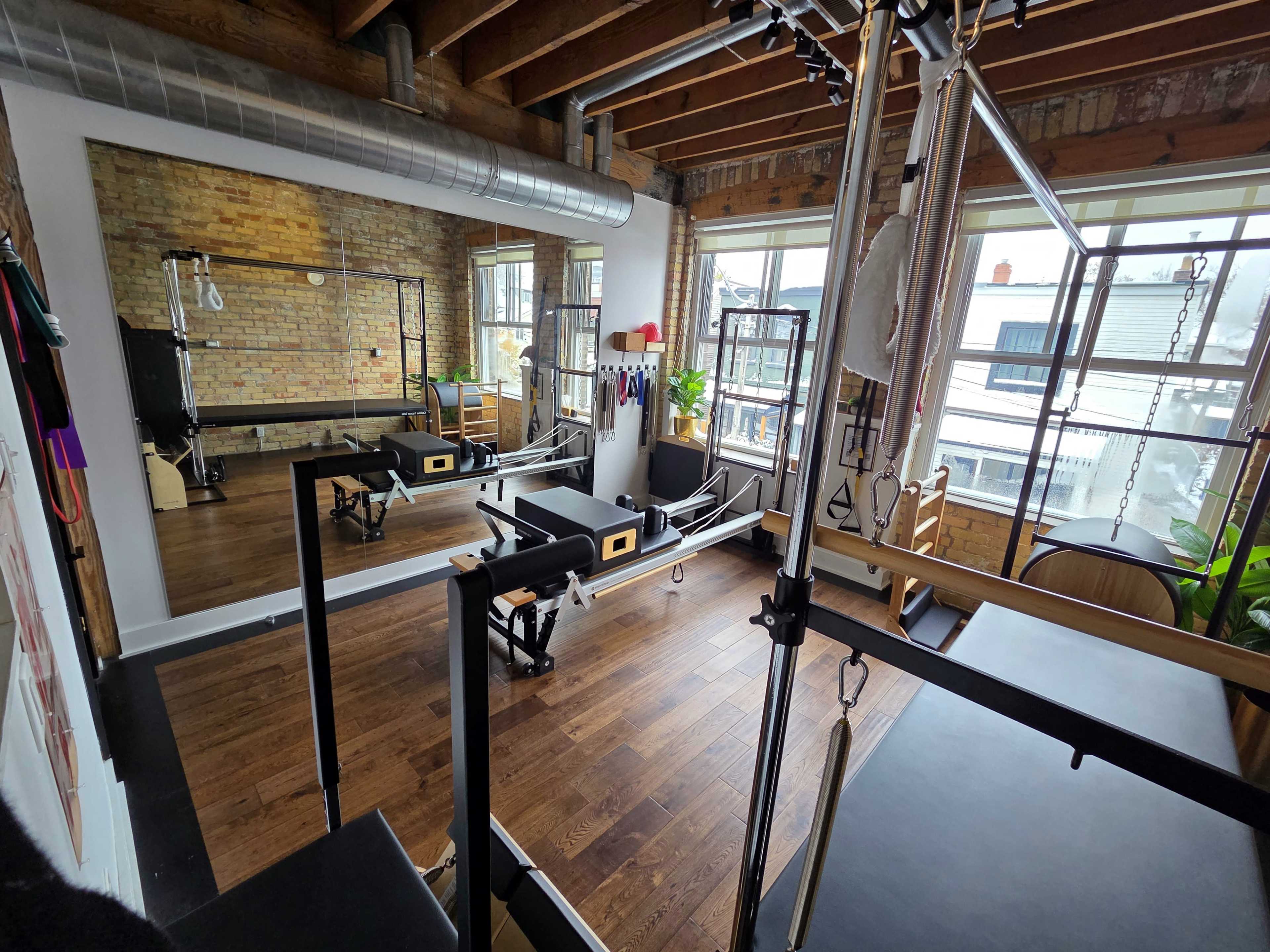 The image shows a well-lit Pilates studio featuring exercise equipment, wooden floors, and large windows.
