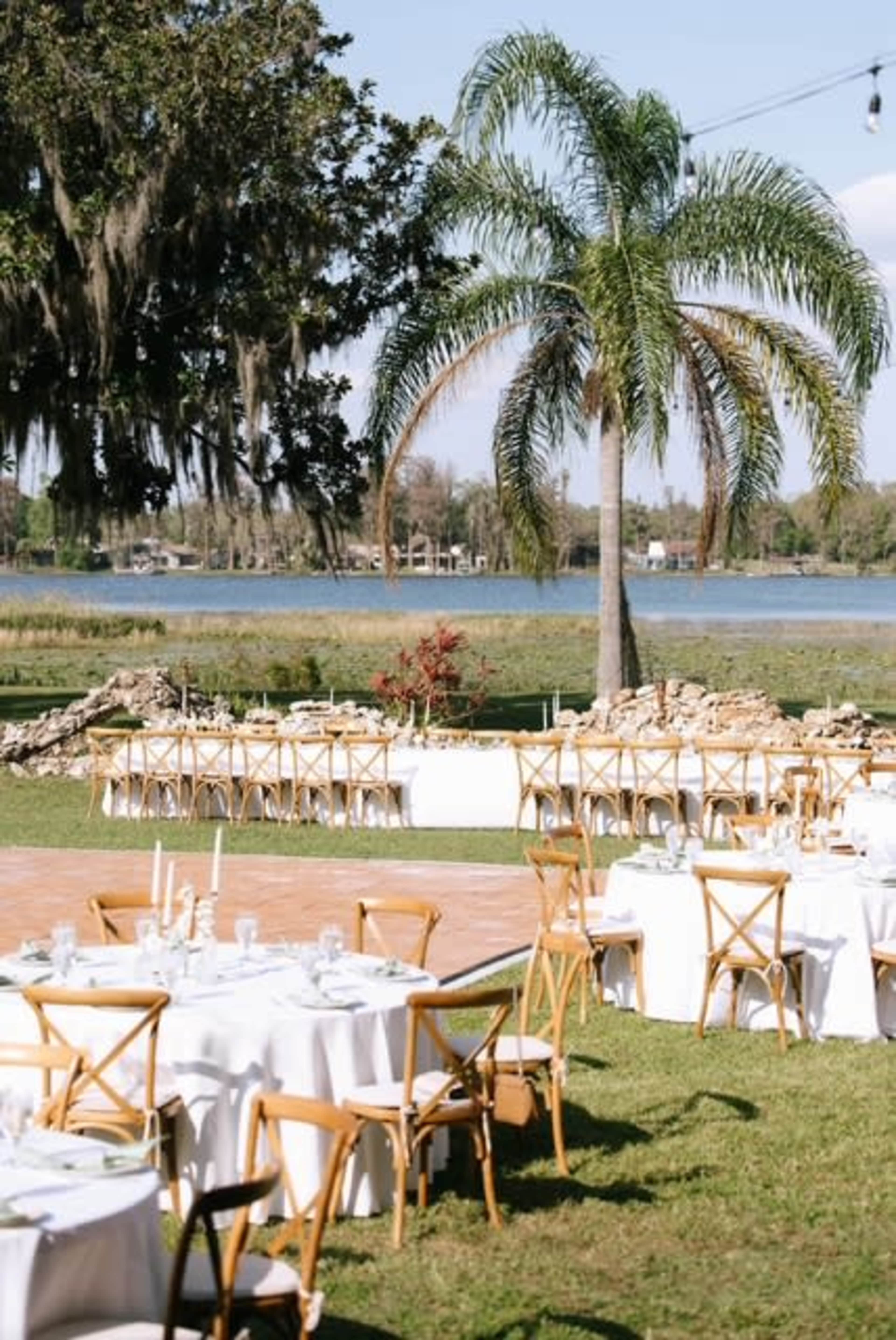 A lakeside event setup features rows of round tables with white tablecloths, wooden crossback chairs, and a backdrop of palm trees and water.
