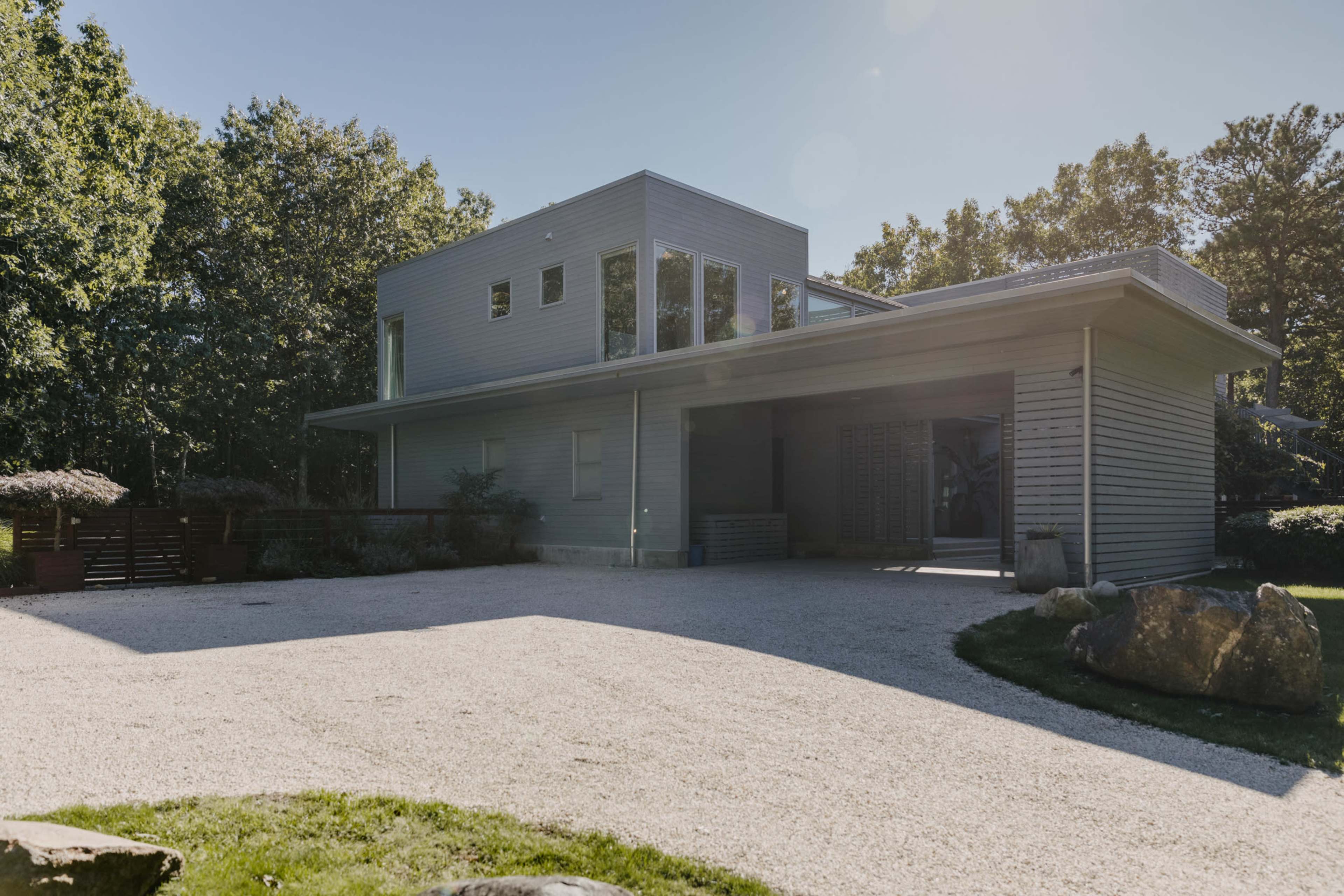 A modern two-story house with large windows and a gravel driveway is surrounded by greenery.