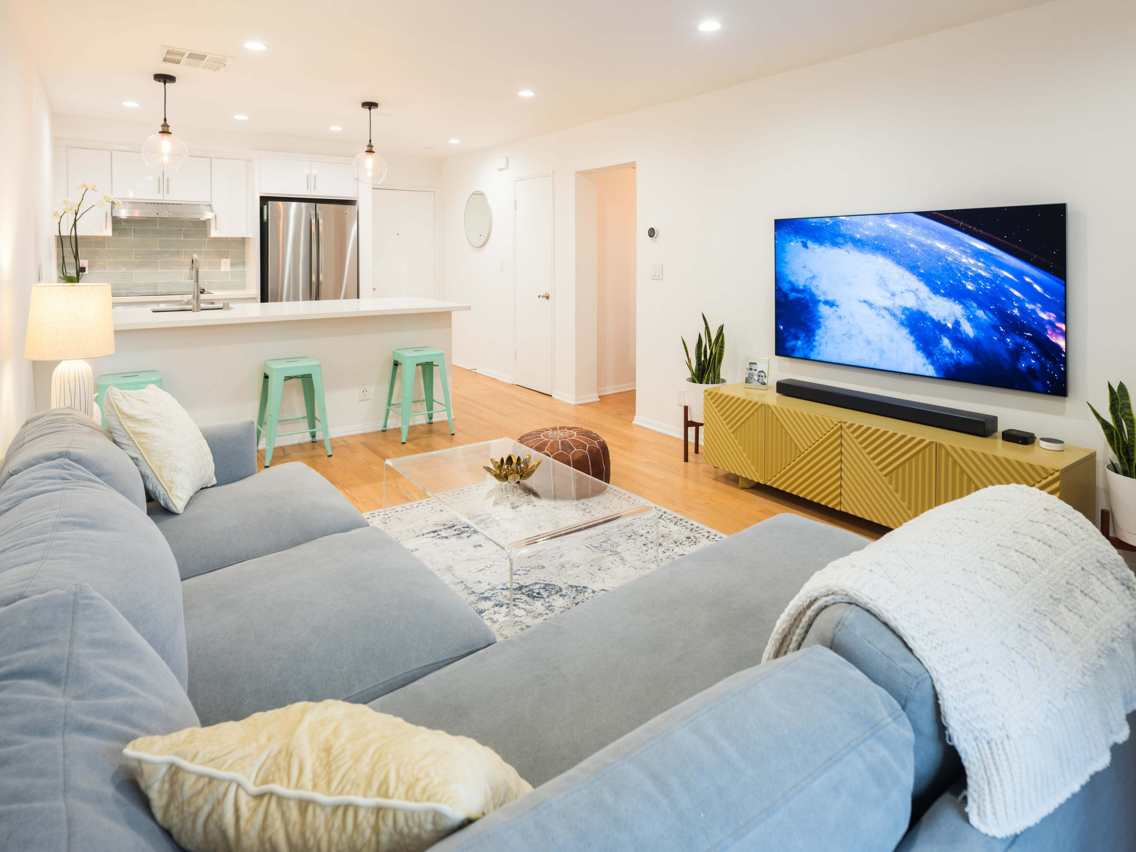 The image shows a modern living room with a gray sectional sofa, a glass coffee table, a large television mounted on a yellow media console, and a bright kitchen area in the background.