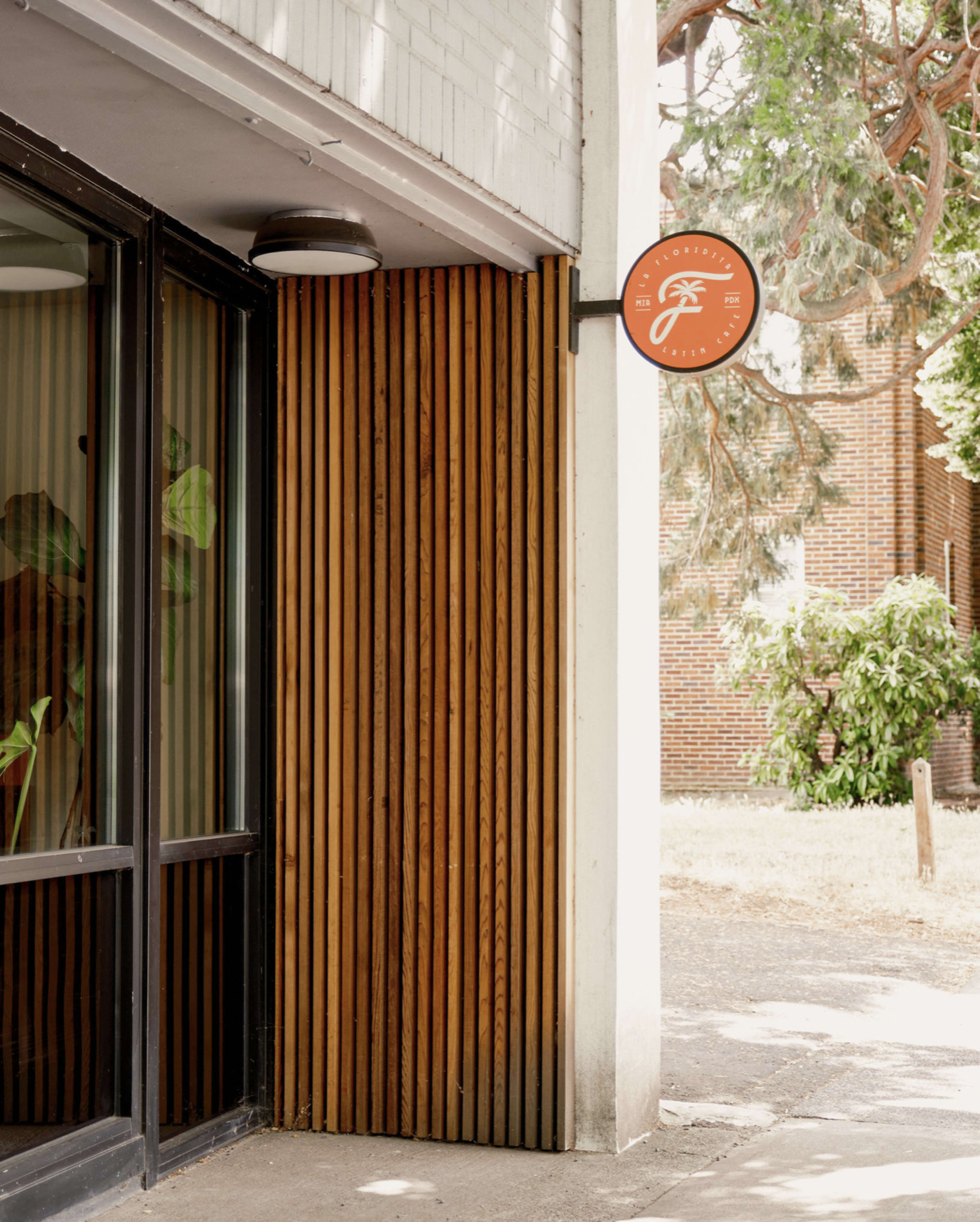 The image shows a building corner with wooden slats, a round sign featuring a logo, and a green plant visible through the glass door.