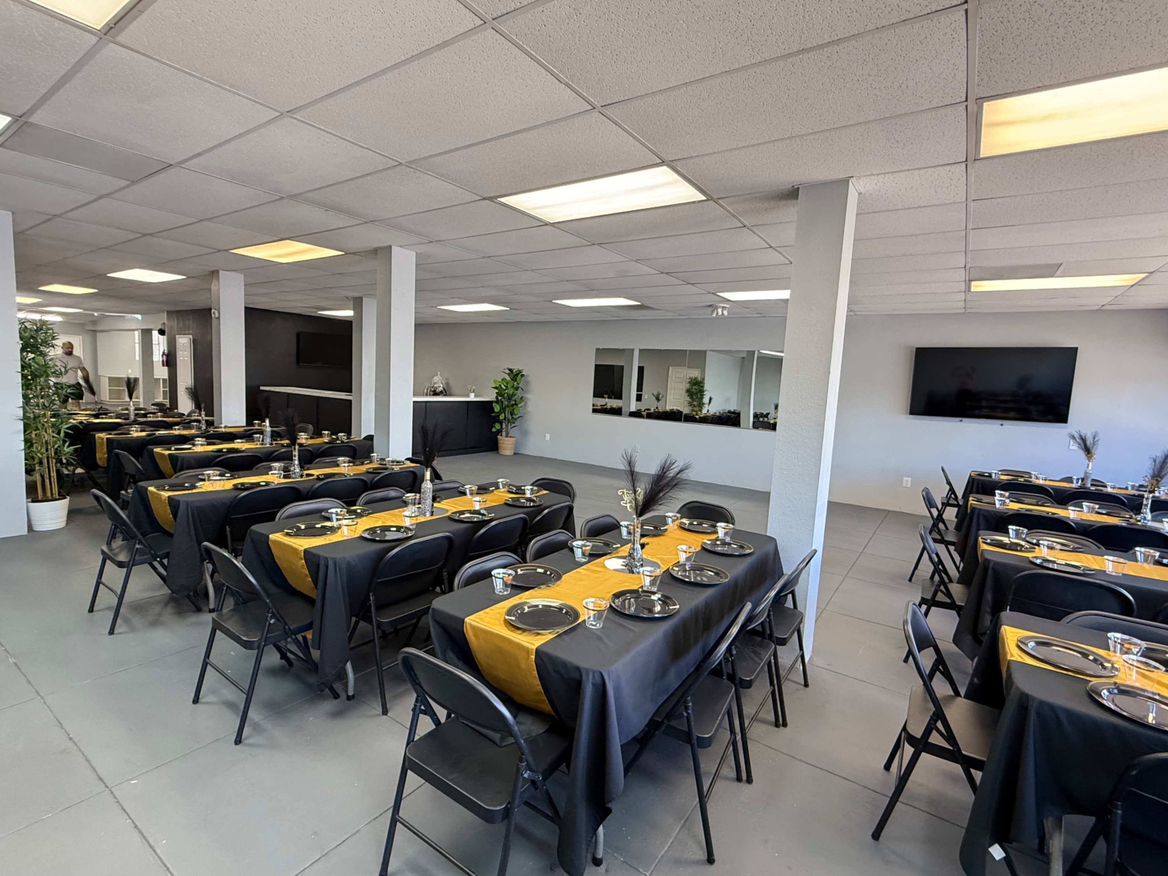 A spacious event room features multiple long tables covered with black and gold tablecloths, each set with plates and utensils.