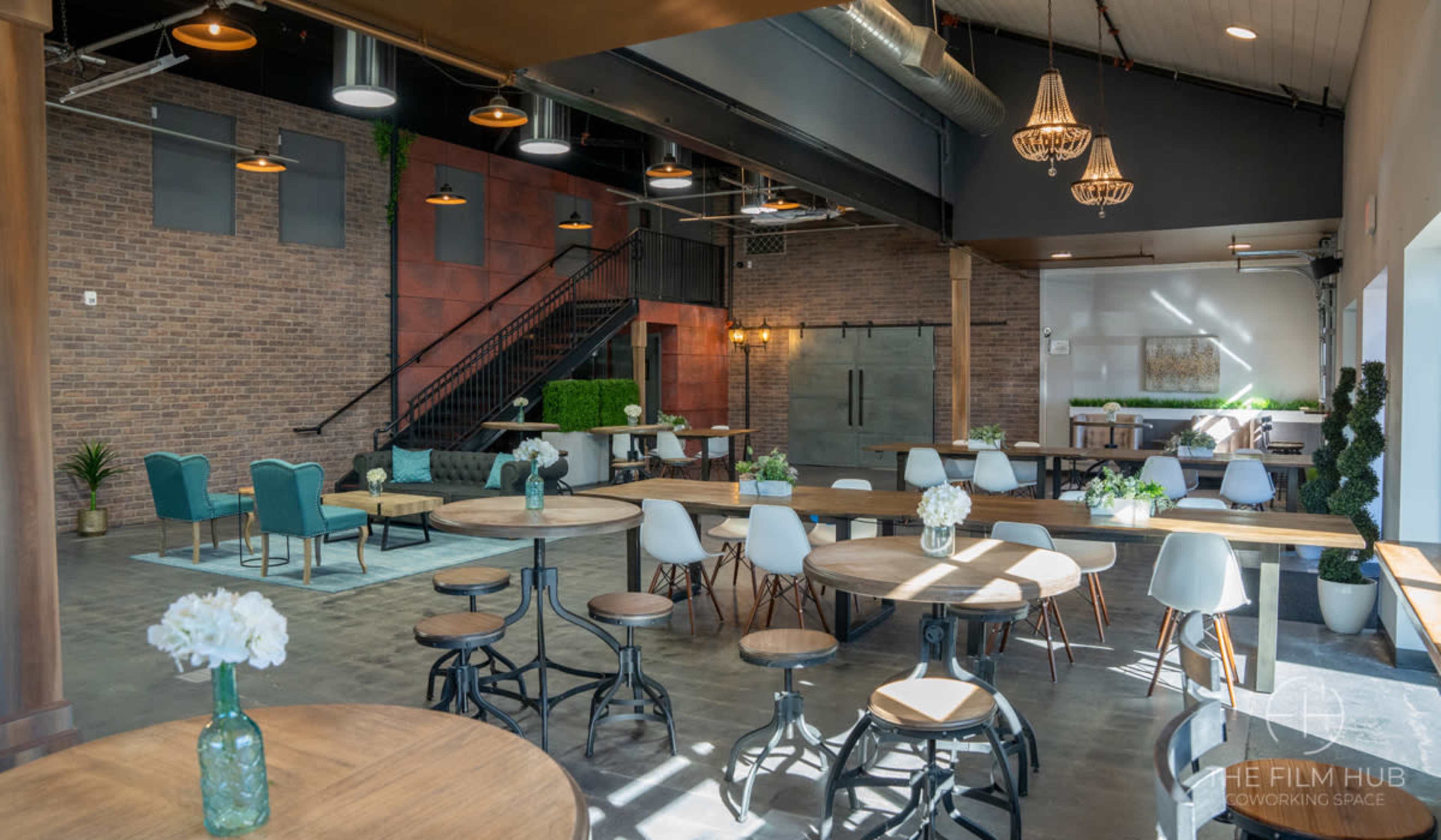 A modern coworking space with wooden tables, green plants, and a staircase in a well-lit interior featuring brick walls and industrial design elements.