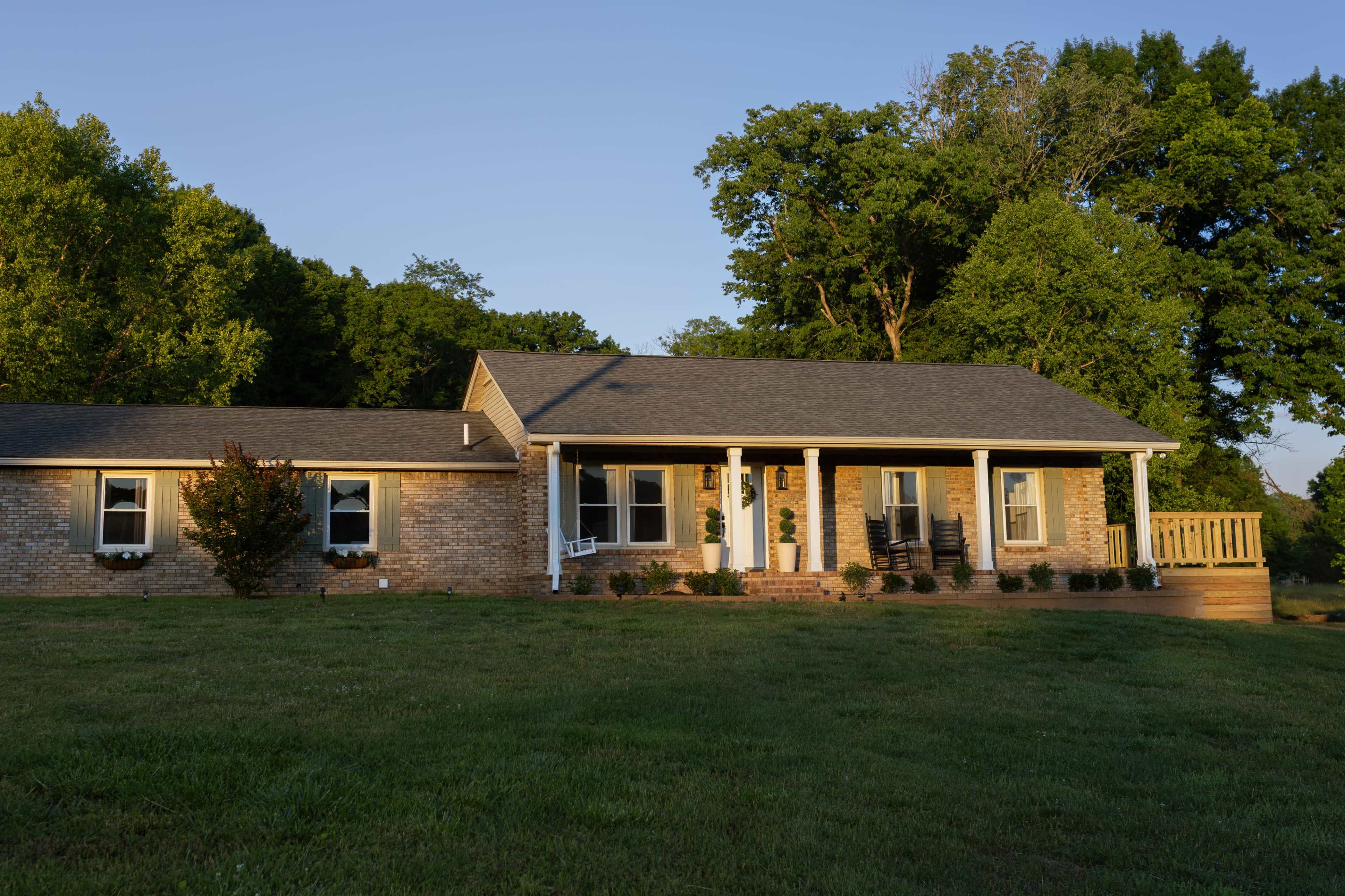 The image shows a single-story brick house with a porch, situated on a grassy lawn surrounded by trees.
