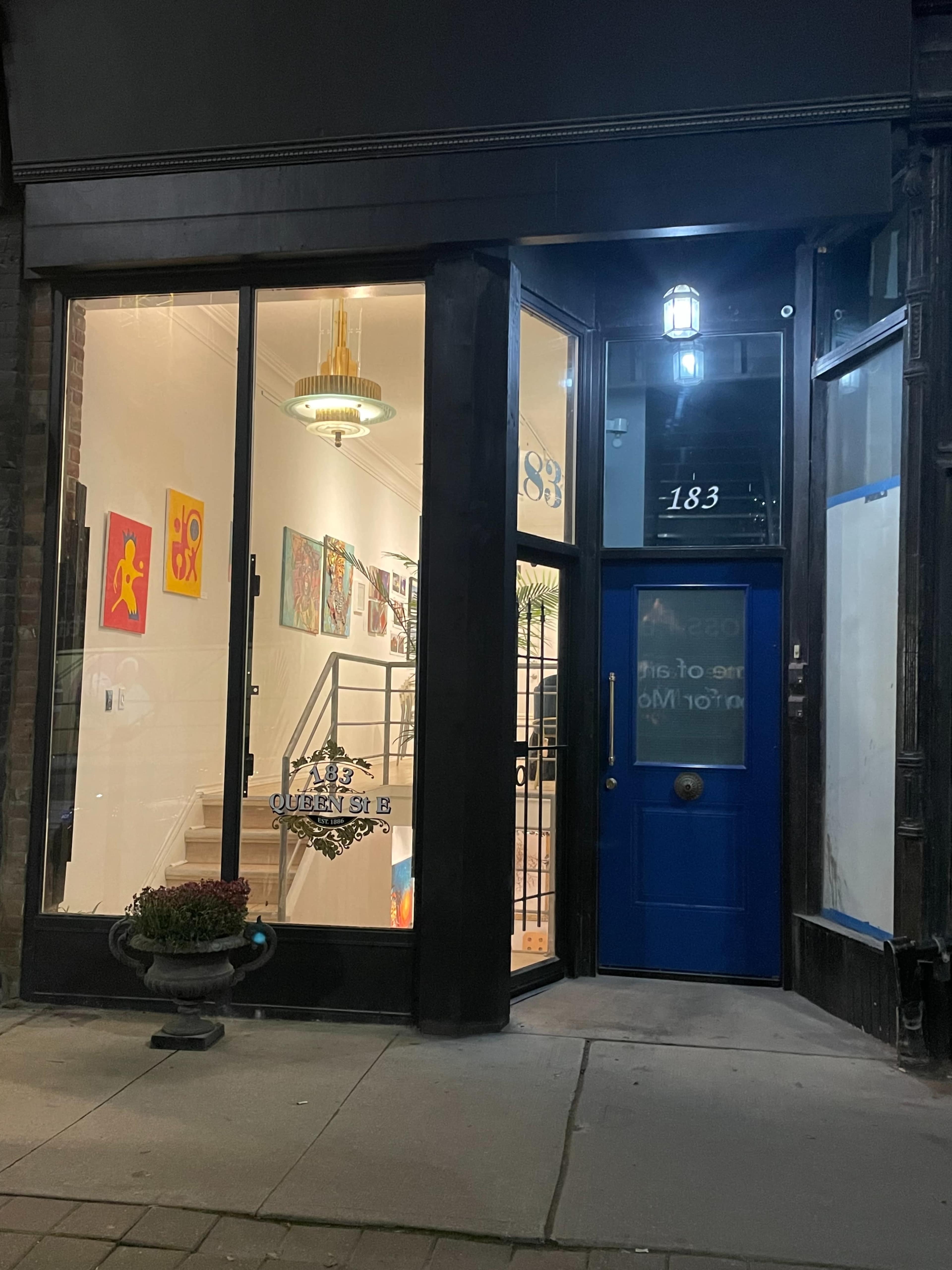 The image shows a storefront with a blue door and large glass windows displaying colorful artwork inside.