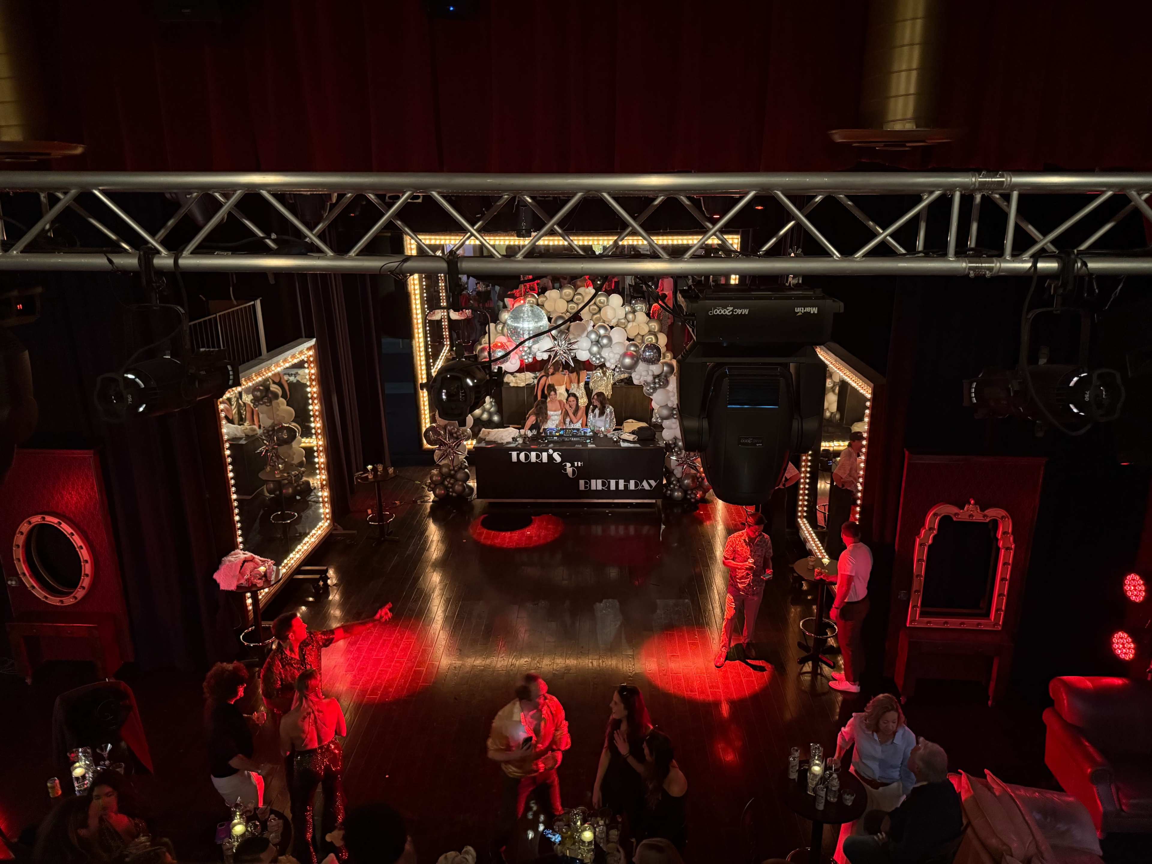 The image shows a lively event space with a decorated table at the center, surrounded by guests and illuminated by bright lights.