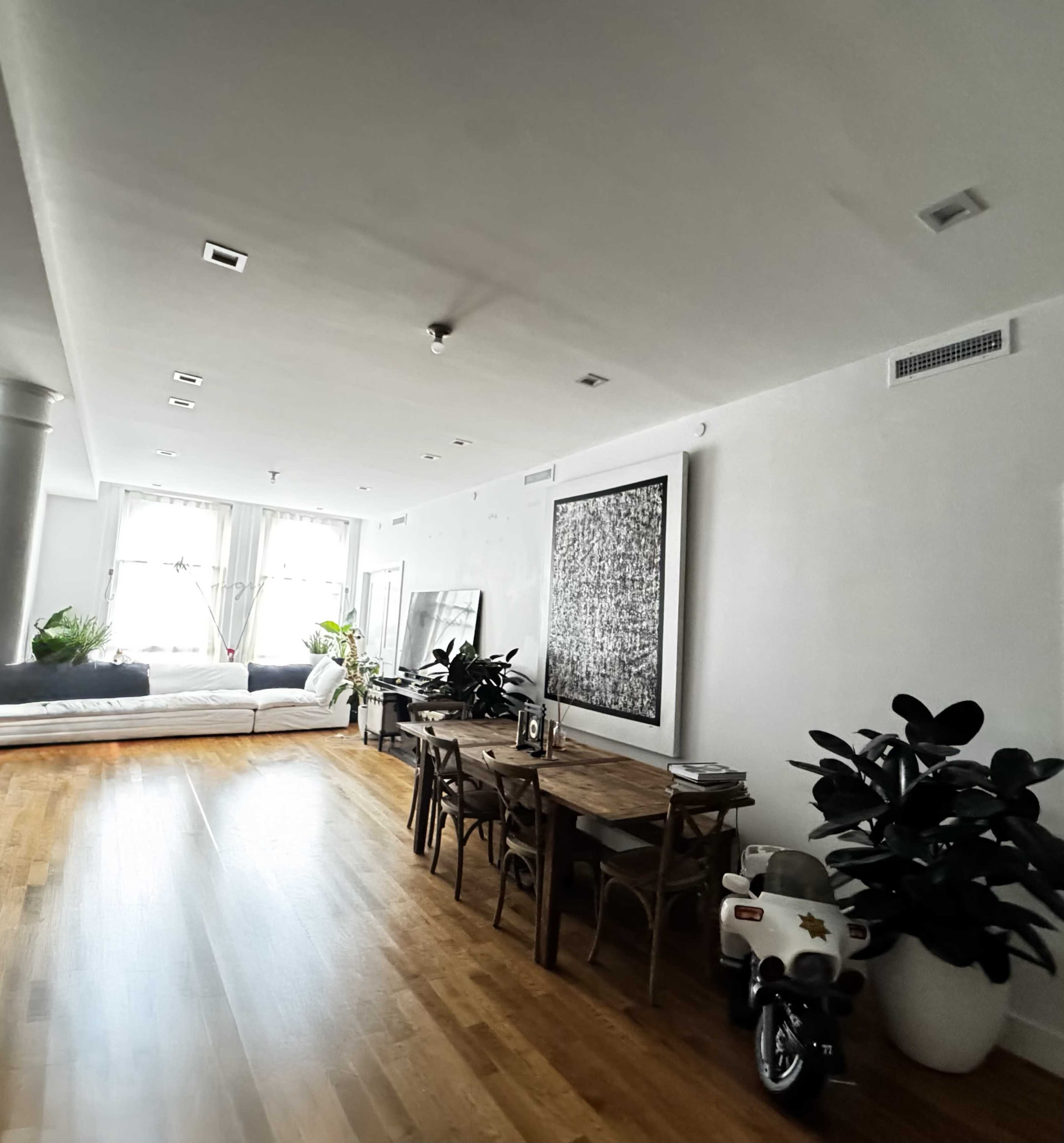 The image shows a bright, modern living space with a dining table, chairs, and plants, beside a bed in a separate area.