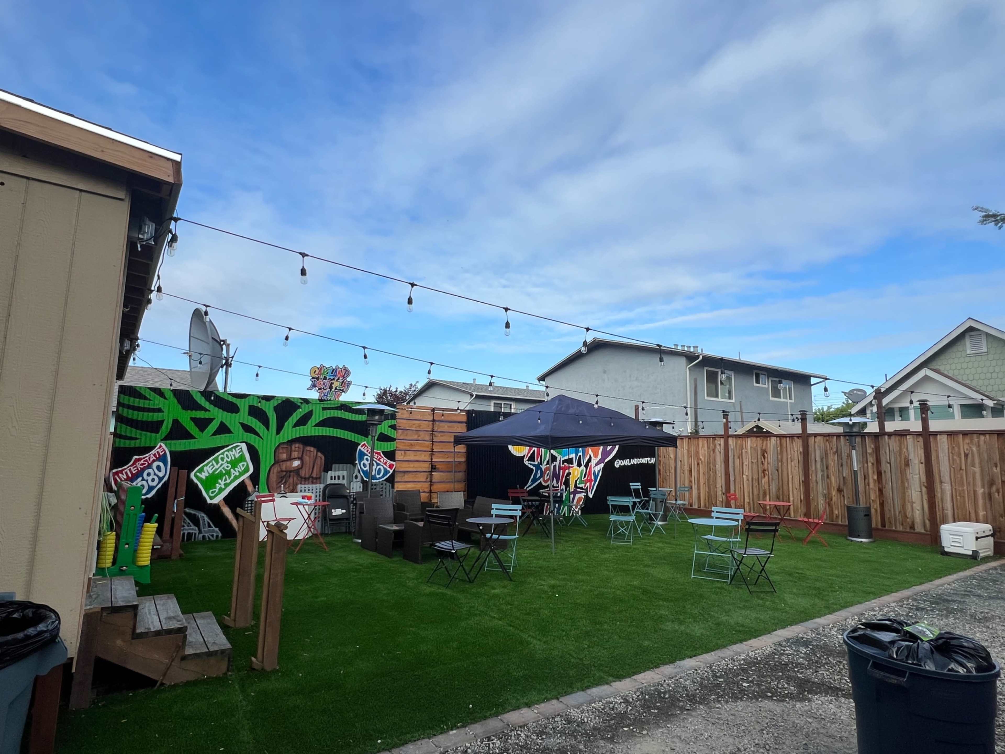 The image shows a backyard area with green artificial grass, outdoor furniture arranged around a tent, and decorative mural elements on the walls.