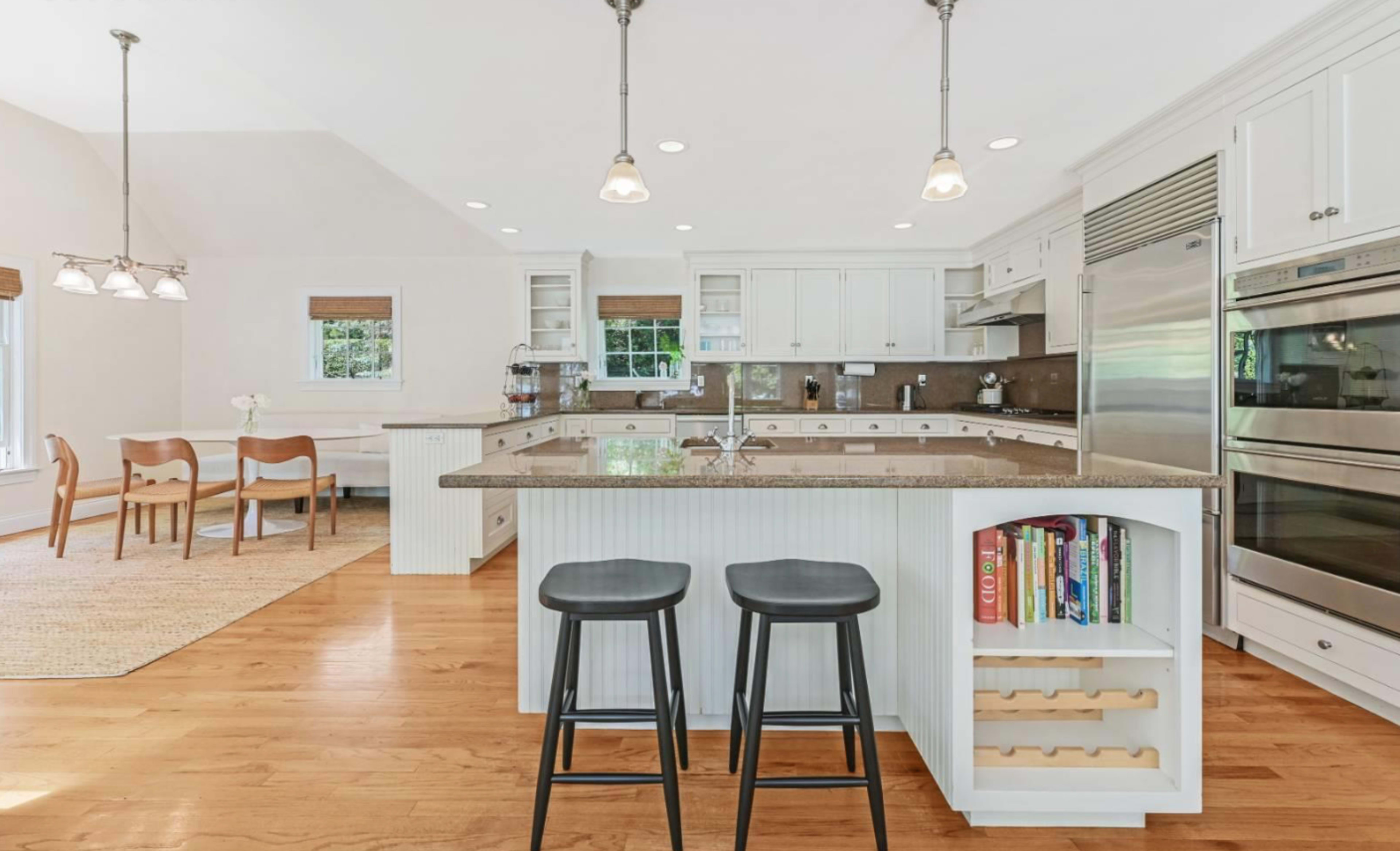 A spacious kitchen features an island with two black stools, stainless steel appliances, and a dining area with wooden chairs.