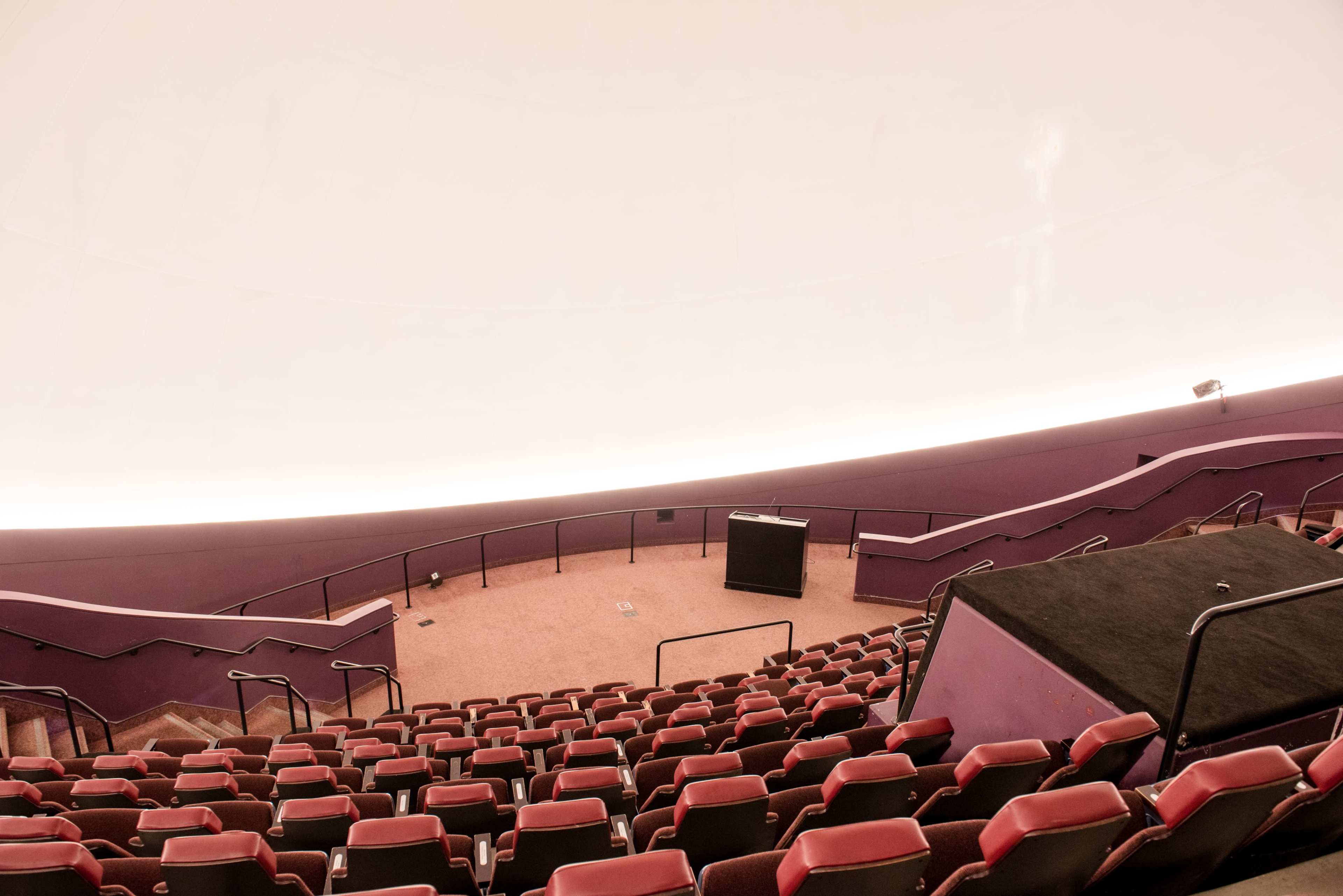 The image shows a spacious, circular auditorium with red seating arranged in a tiered layout, leading up to a curved white ceiling.