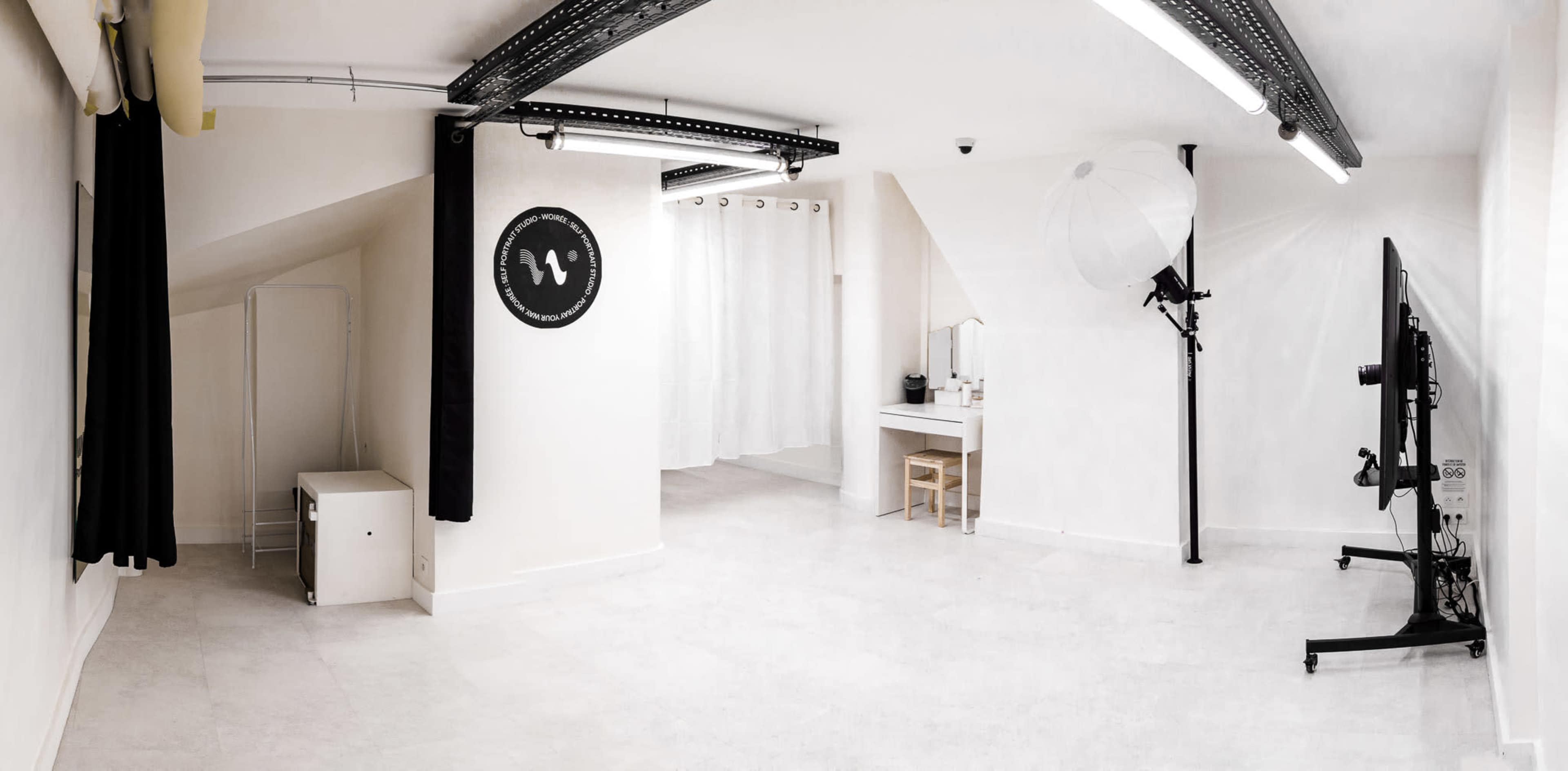 The image shows a spacious, minimalist studio with white walls, a small white chair and table, a standing light source, and curtains on one side.