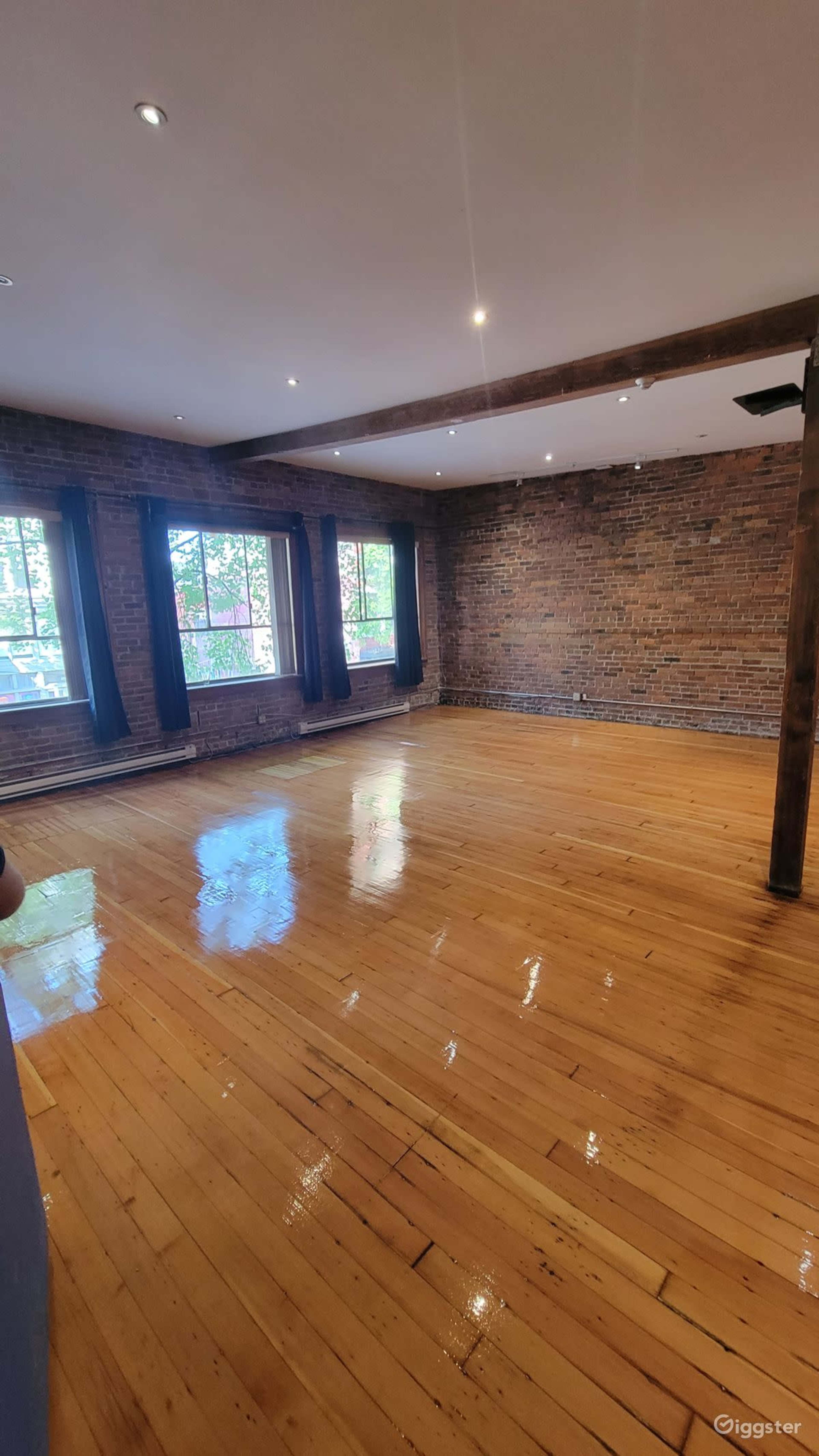 The image shows a spacious, empty room with hardwood floors, exposed brick walls, and large windows allowing natural light to enter.