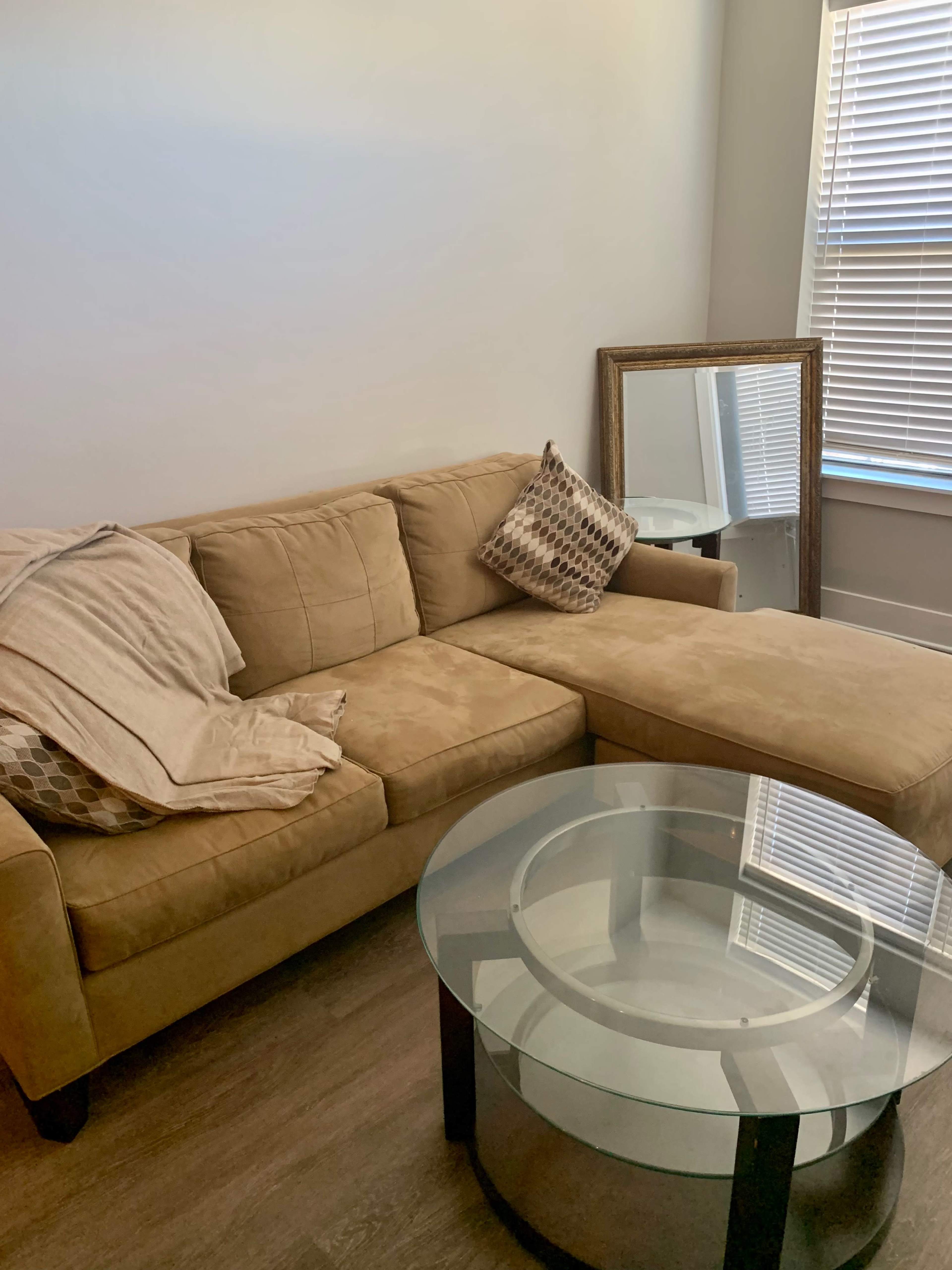 A tan sectional sofa with a decorative pillow is positioned next to a round glass coffee table and a mirror, under a window with blinds.