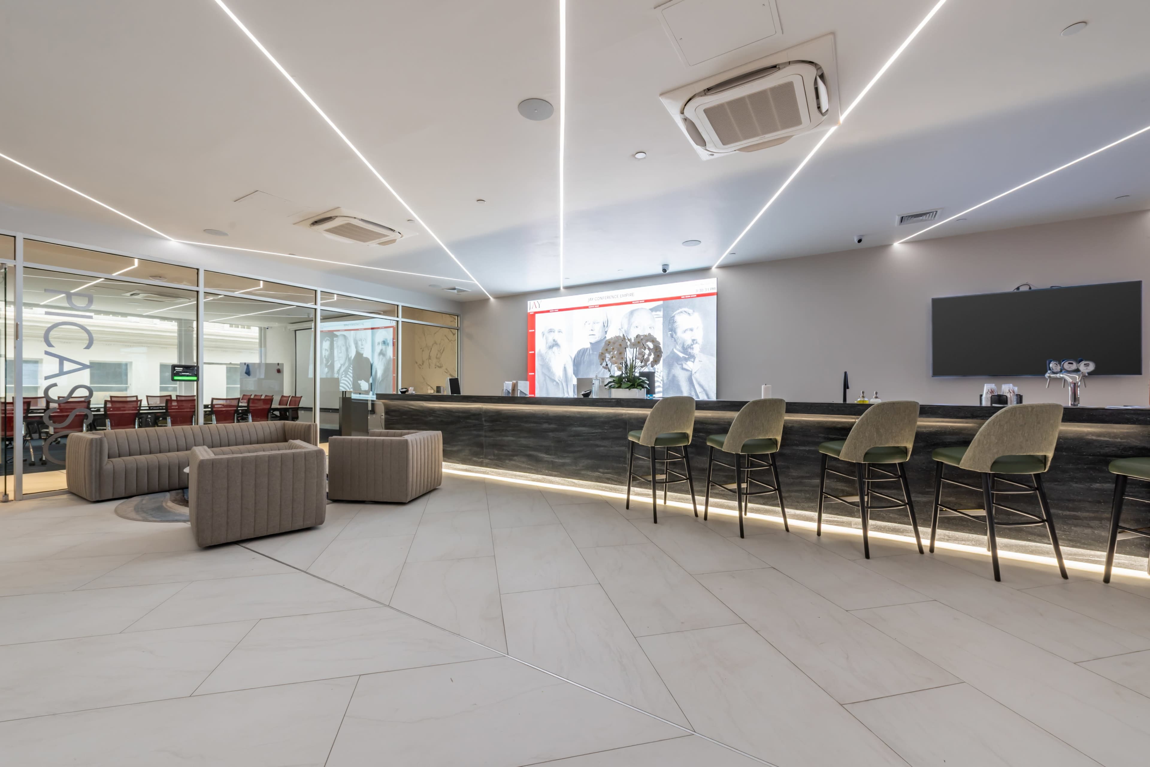 The reception area features a modern bar counter with high stools and a seating arrangement of low, curved sofas, set against a backdrop of large glass windows and a digital display.