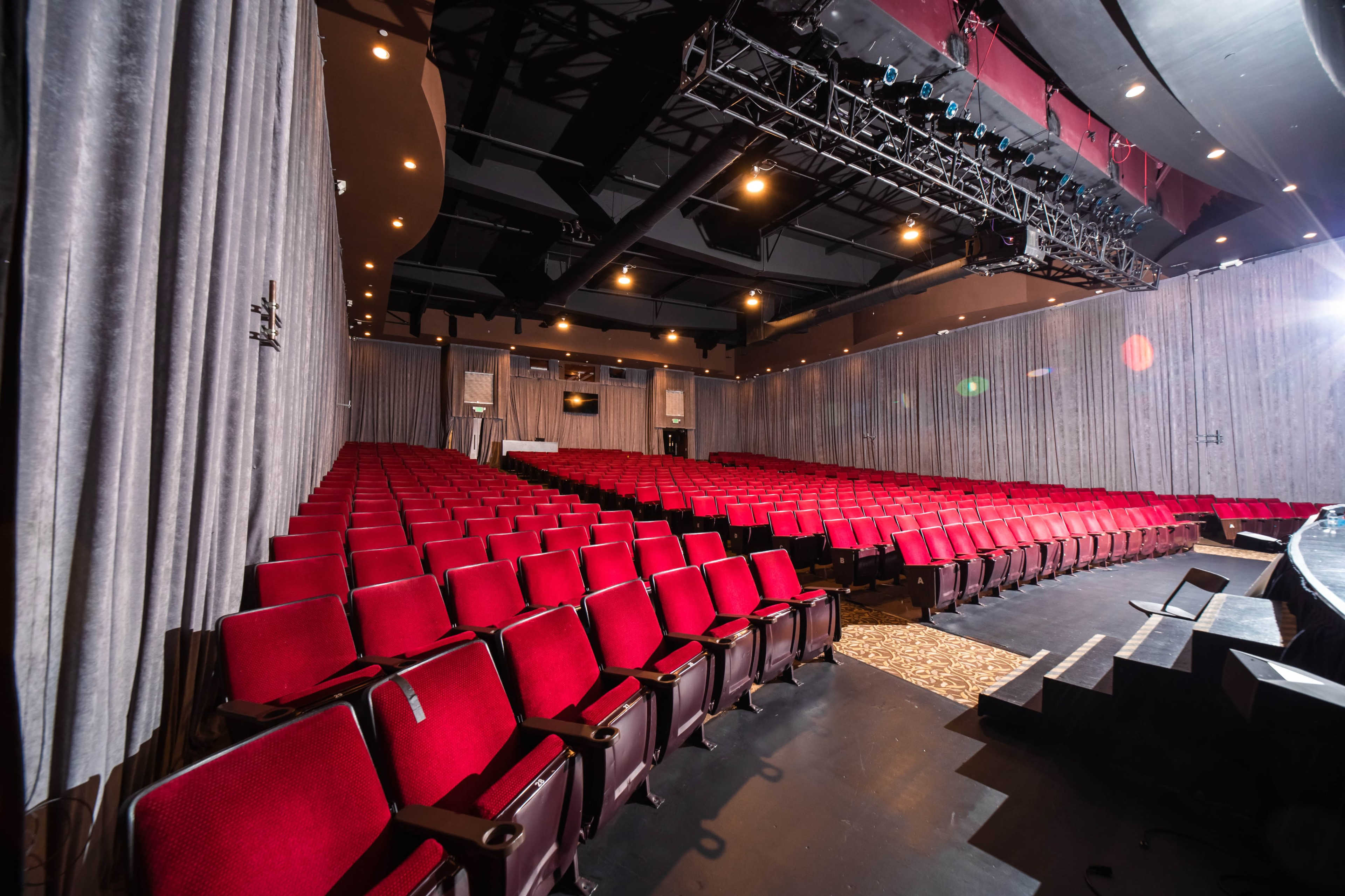 A well-equipped theater features rows of red seats facing a stage, with curtains and overhead lighting in the background.