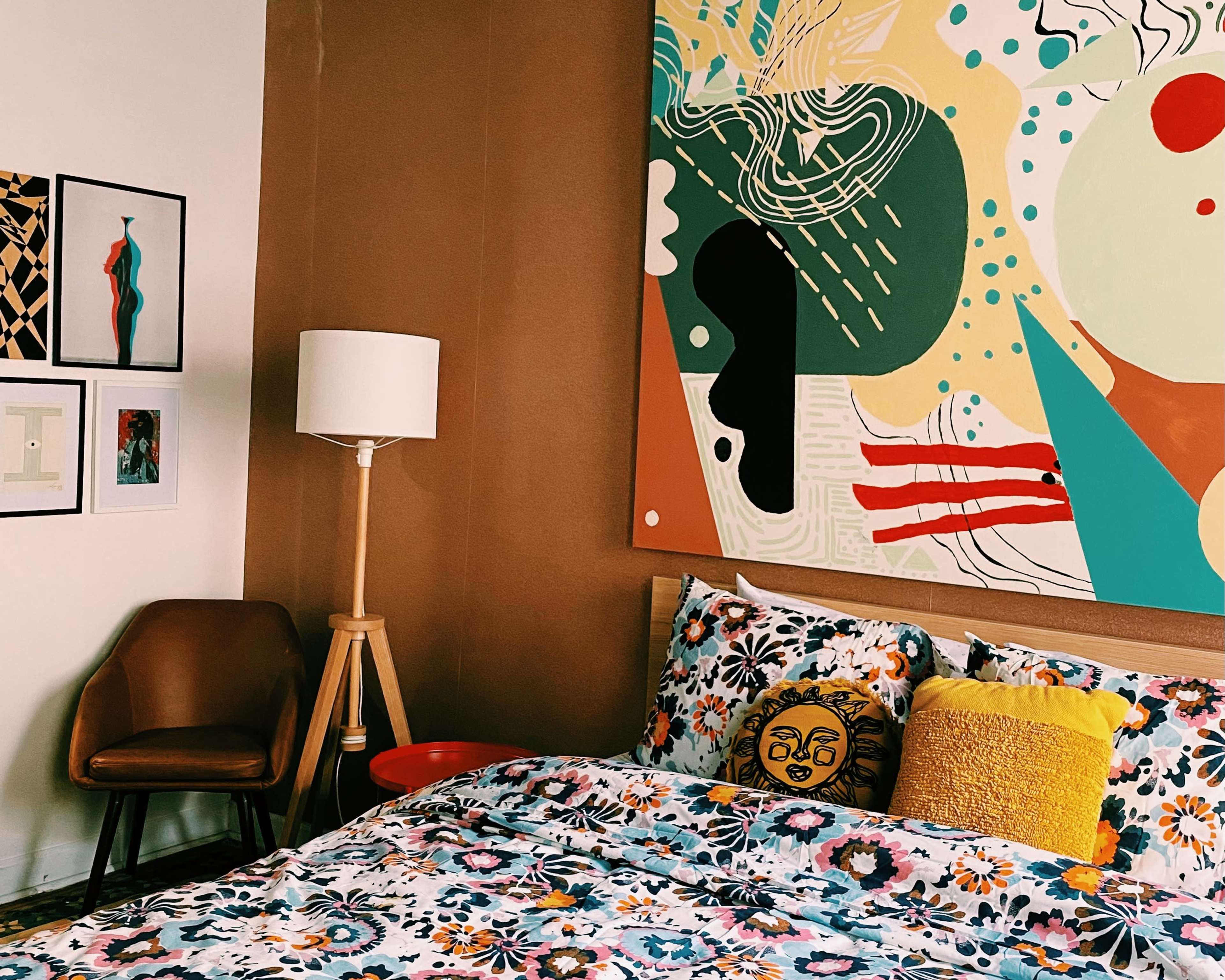 The image shows a cozy bedroom with a colorful bedspread, decorative pillows, a standing lamp, and an abstract painting on the wall.
