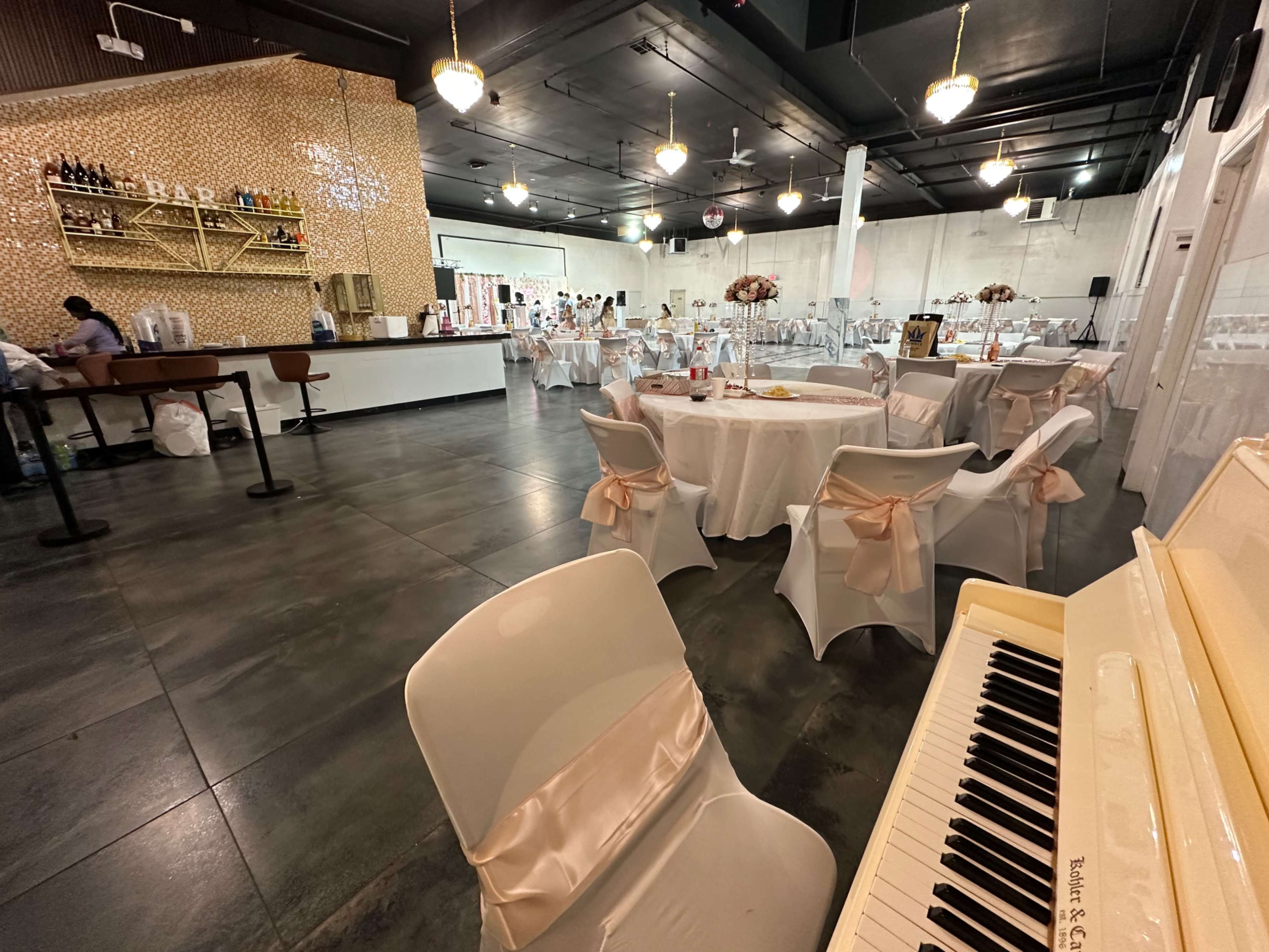 The image shows a spacious event venue with round dining tables elegantly set up, a white piano in the foreground, and a bar area in the background.