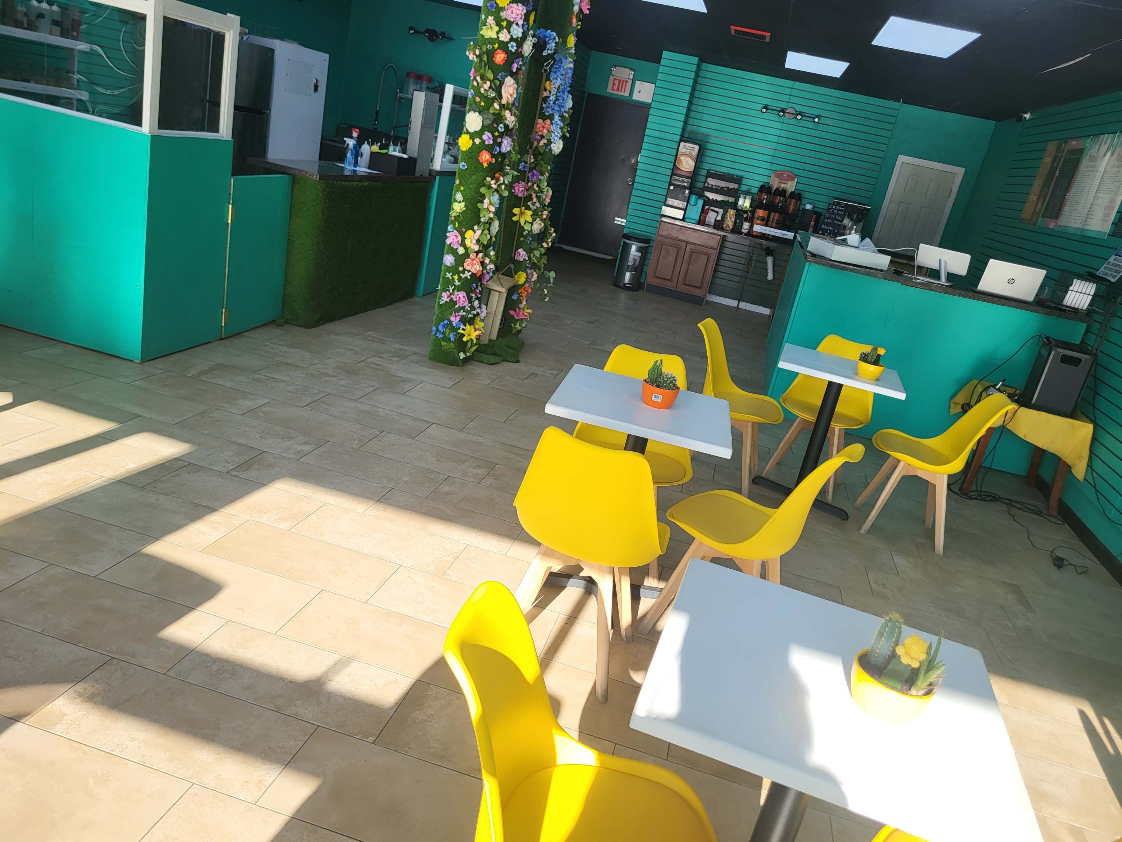 The interior of a colorful café features yellow chairs and tables, a floral decoration on the wall, and a service area in the background.