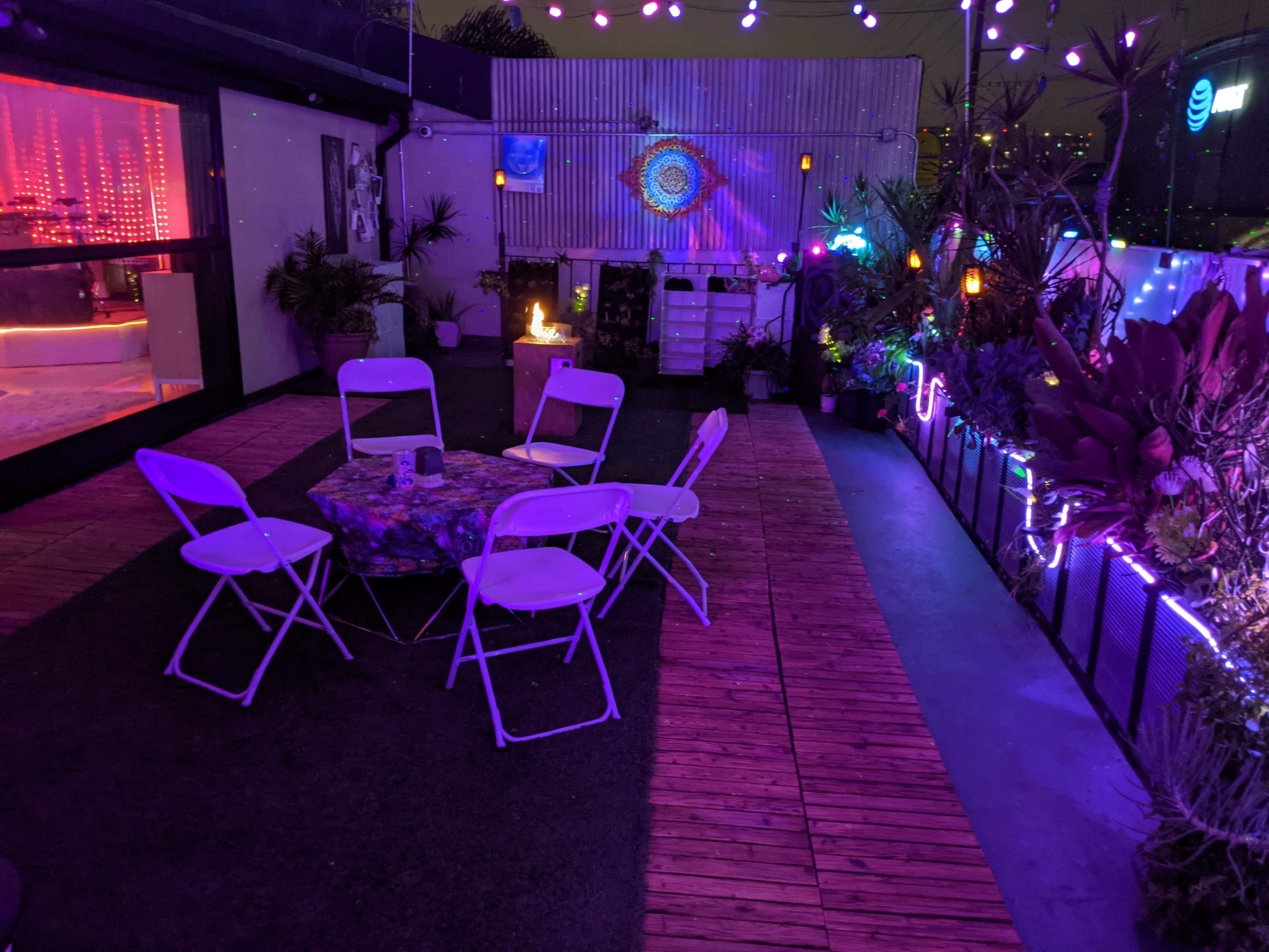 A patio area features a circular arrangement of white folding chairs around a colorful table, surrounded by plants and illuminated by colorful string lights.