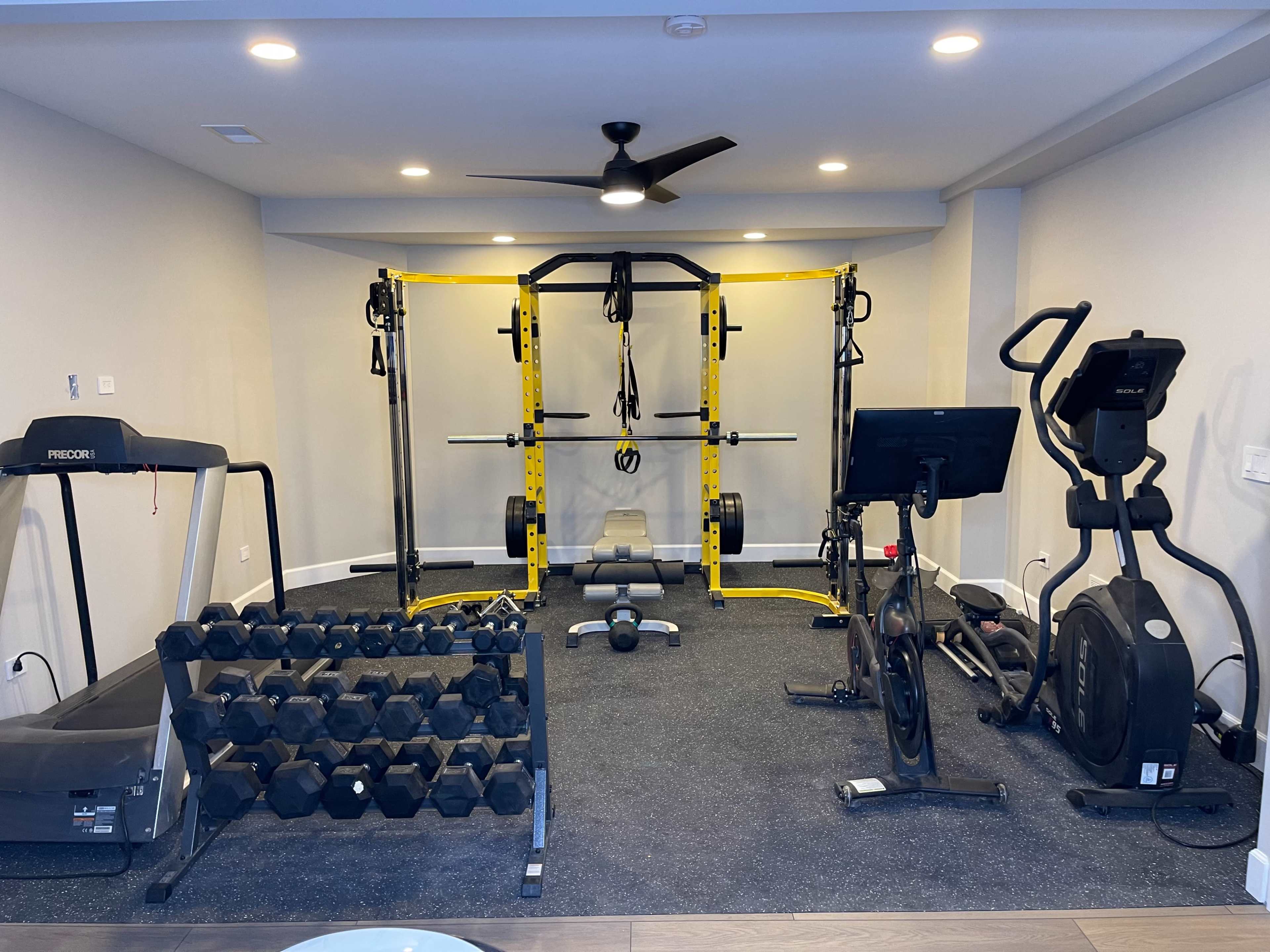 The image shows a home gym equipped with a multi-purpose weight rack, dumbbells, a treadmill, an elliptical machine, and a stationary bike on a rubber mat surface.