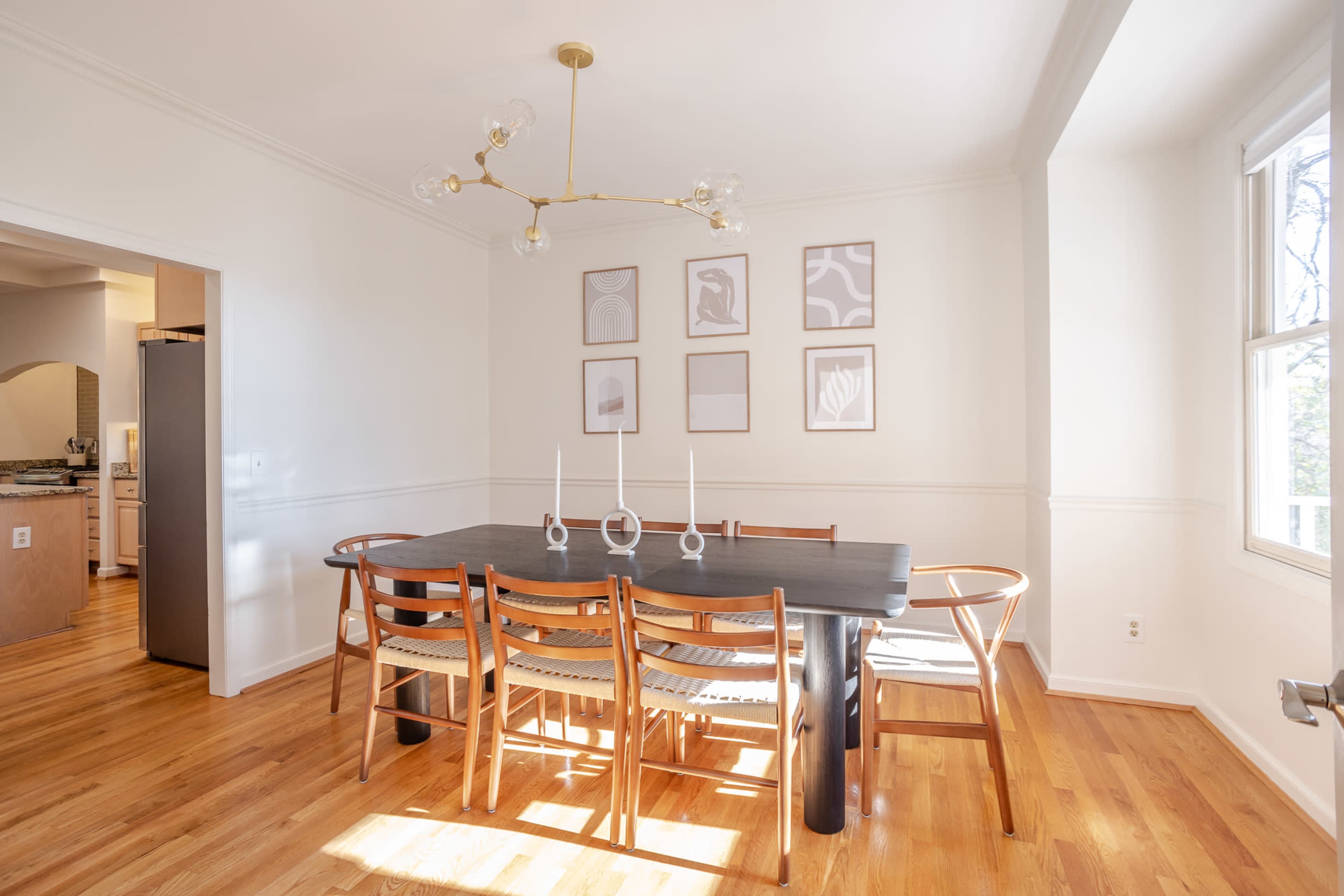 A modern dining room features a large table surrounded by wooden chairs, with framed artwork on the walls and natural light coming from a window.