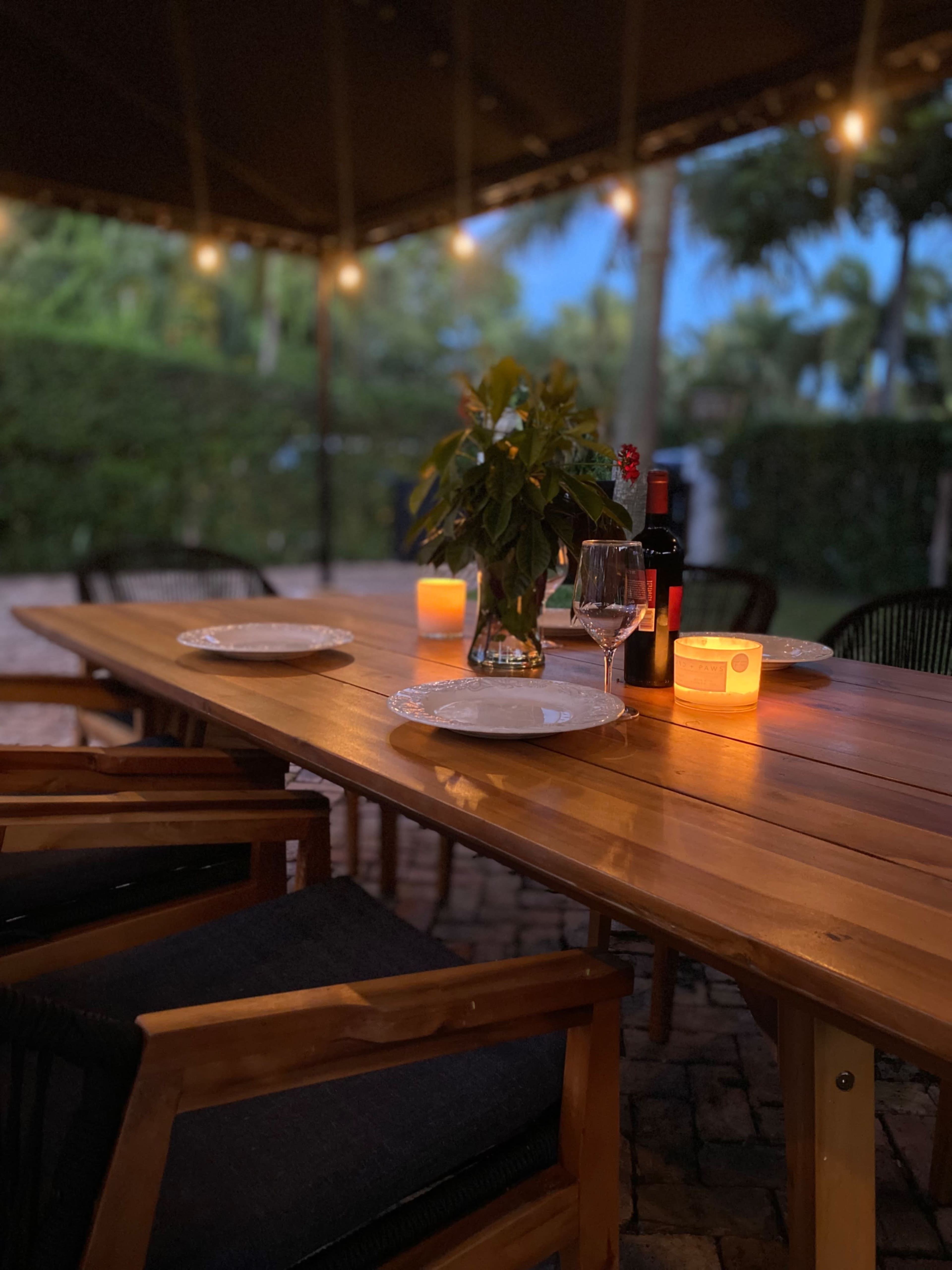 A wooden dining table is set with plates and wine glasses under a canopy illuminated by string lights and candles.