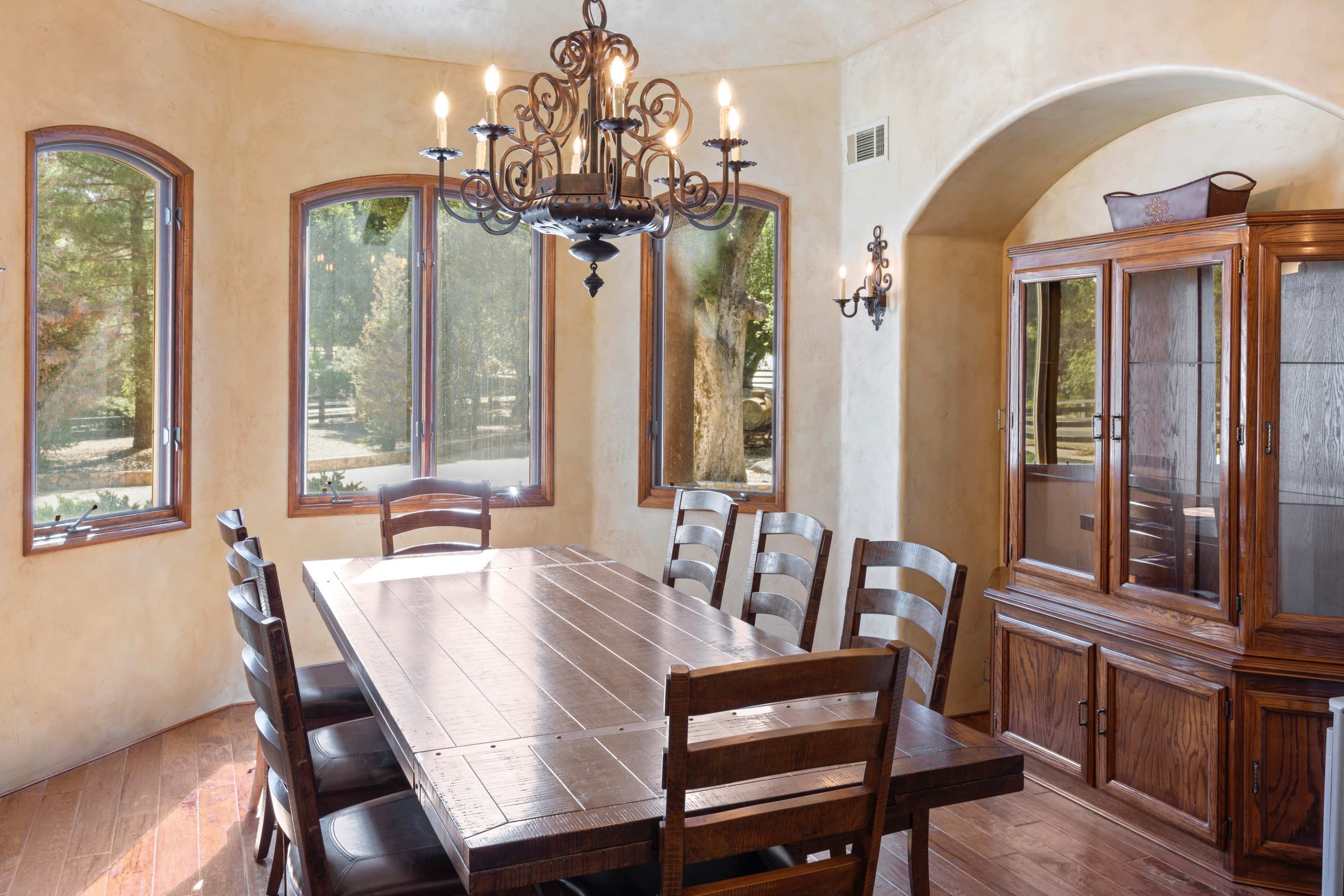 A wooden dining table with eight chairs is set in a well-lit room with large windows and a chandelier.