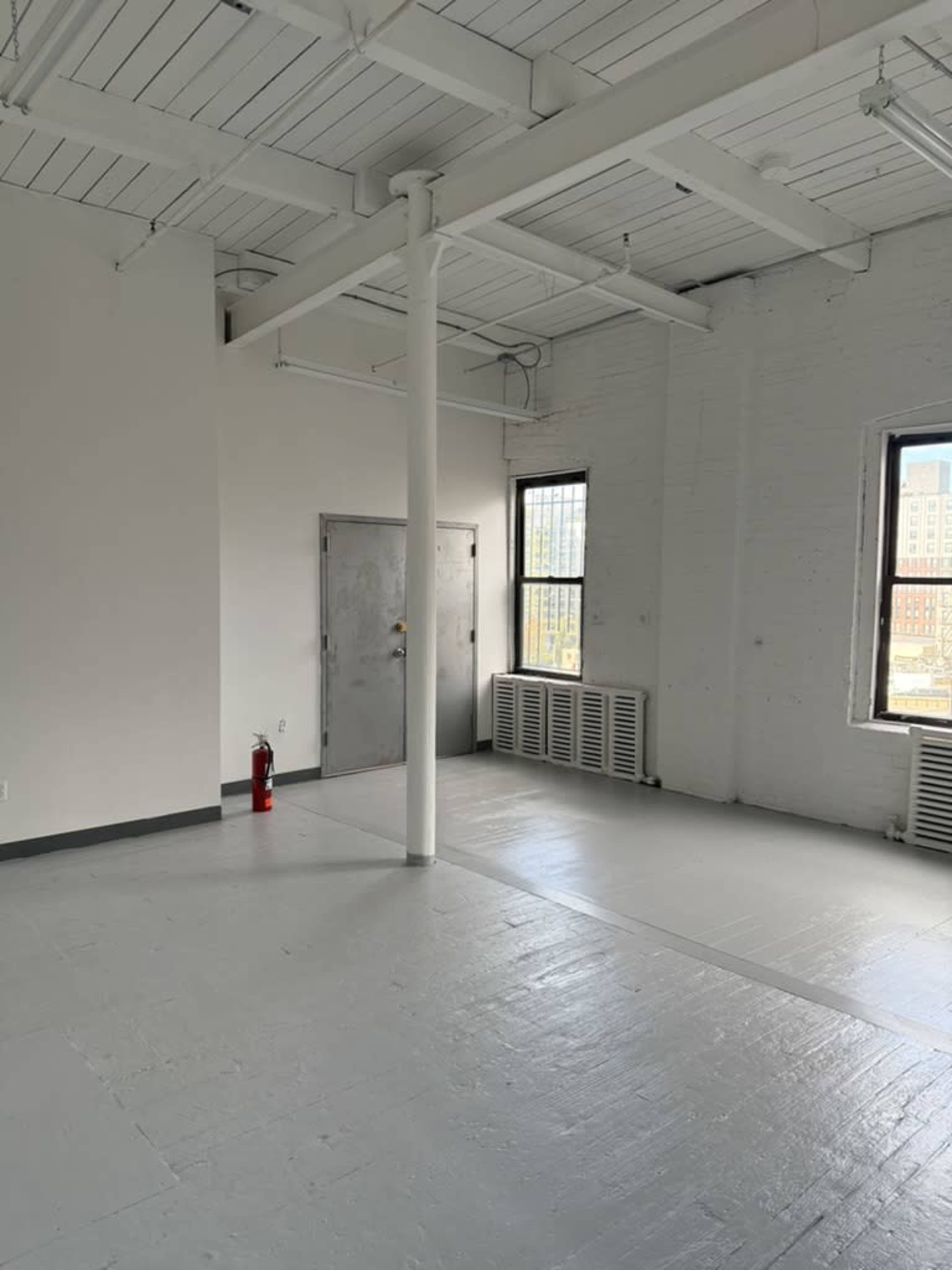 The image shows an empty room with white walls, large windows, a gray floor, a metal door, and a fire extinguisher against the wall.