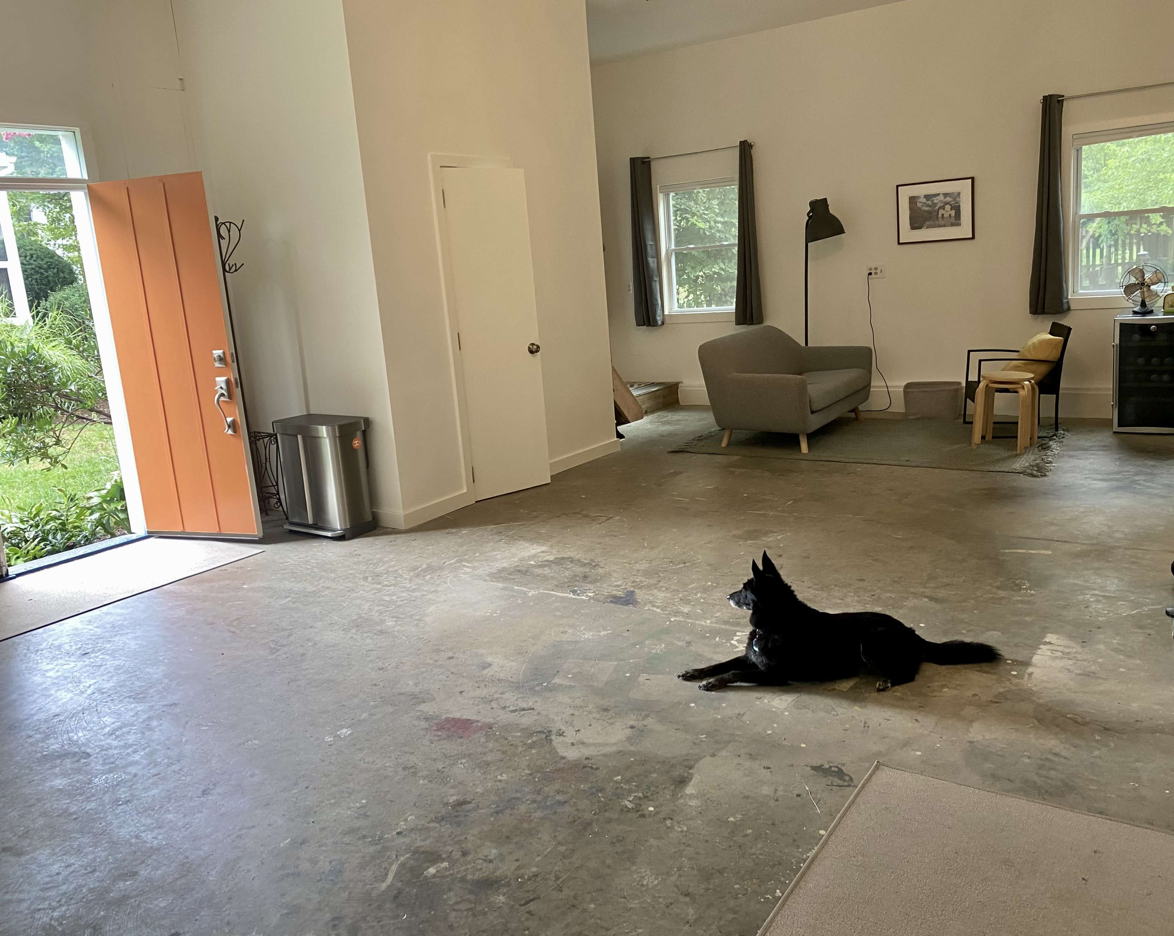 A black dog lies on the concrete floor of an interior space with an open orange door leading to an outdoor area.