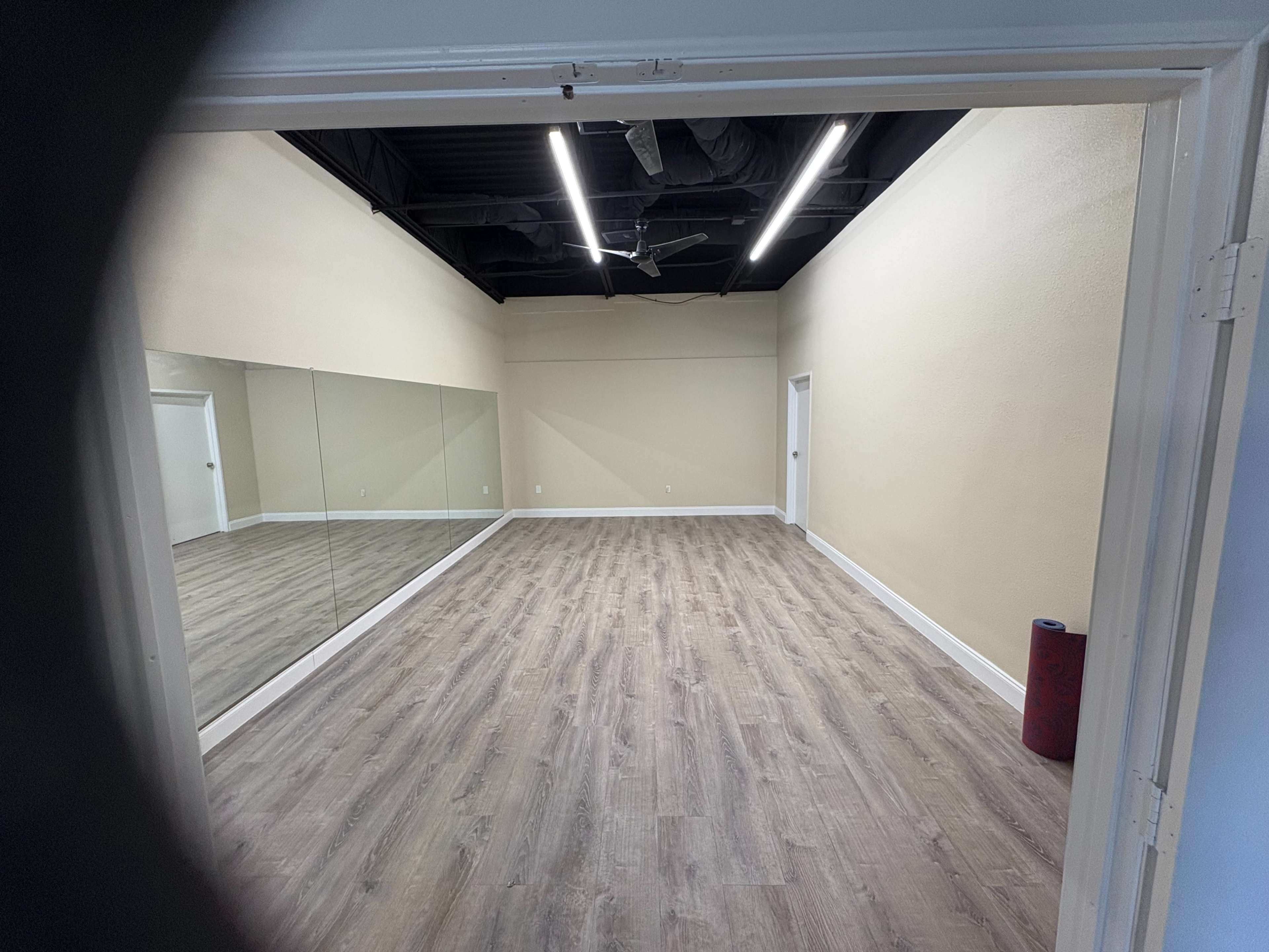 The image shows a spacious, empty room with wooden flooring, a large mirror on one wall, and a door on the opposite side.