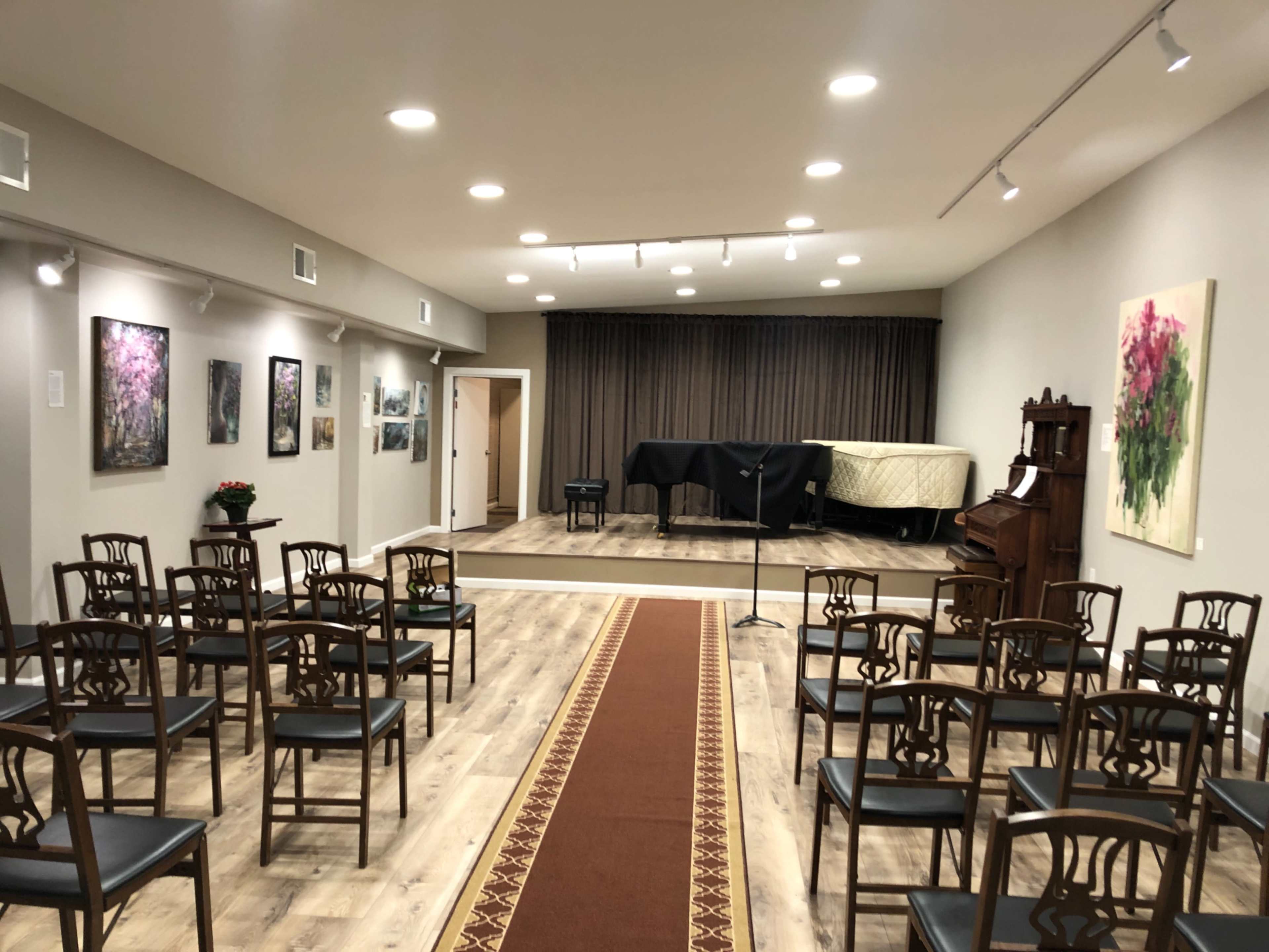 A music performance space with rows of wooden chairs facing a stage covered with a black cloth and a piano, surrounded by framed artwork on the walls.