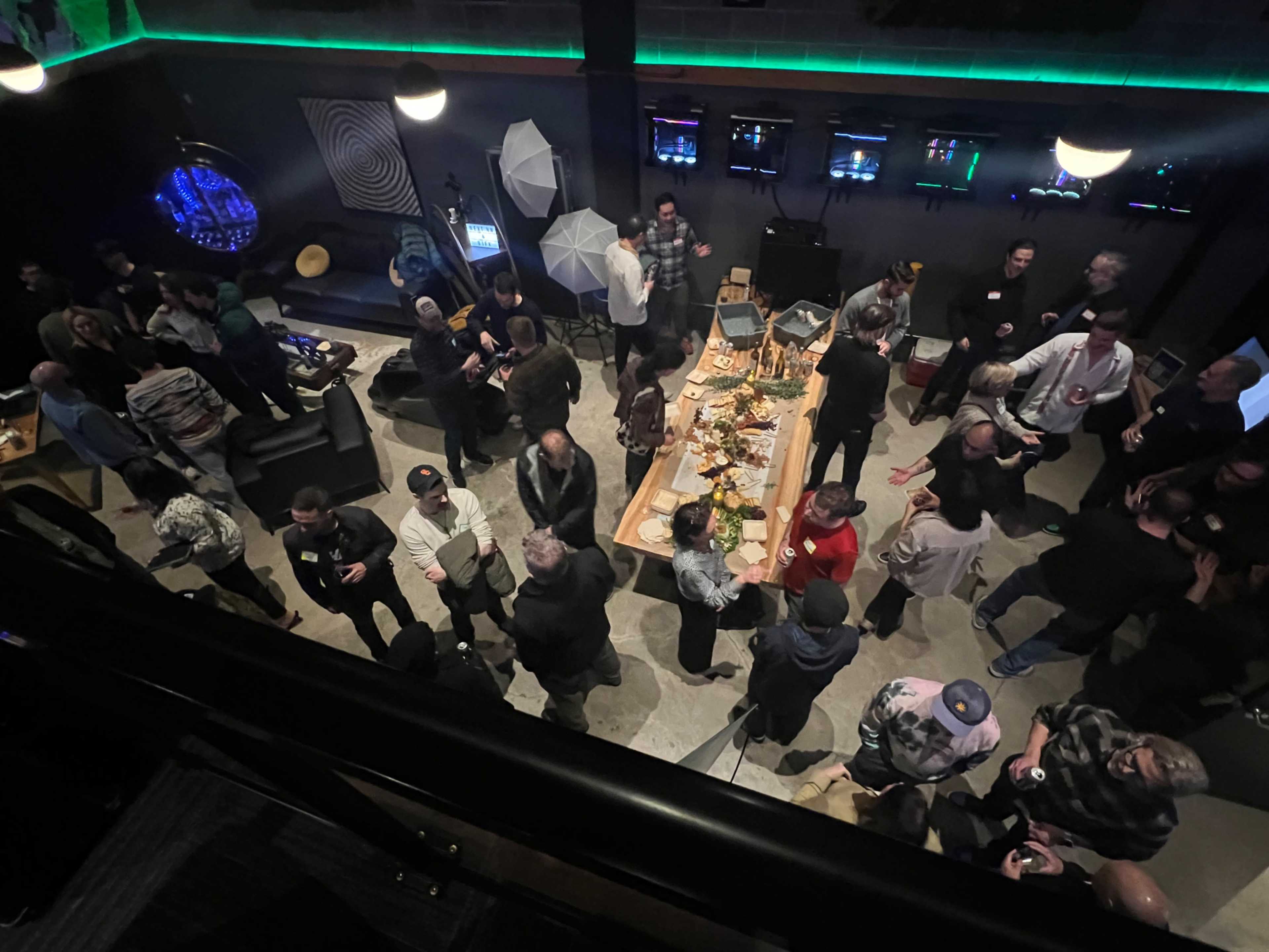 A crowded event space with people mingling around a long table filled with food and drinks, while others stand in conversation.
