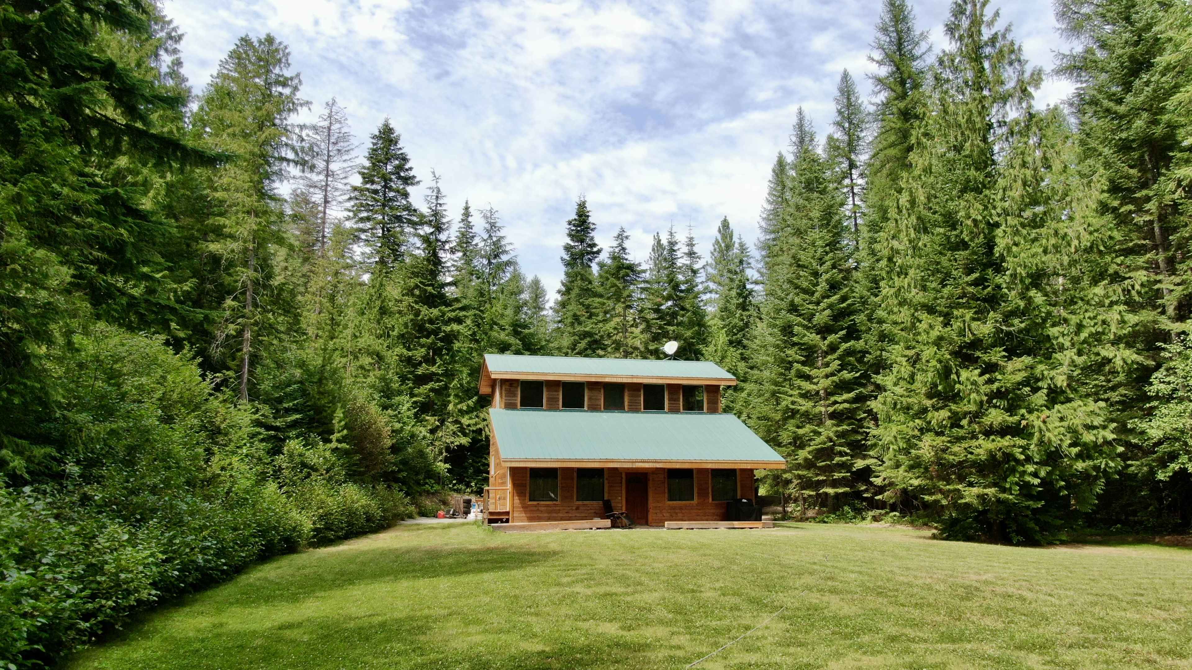 A two-story wooden cabin with a green metal roof is situated in a lush, forested area surrounded by tall pine trees.