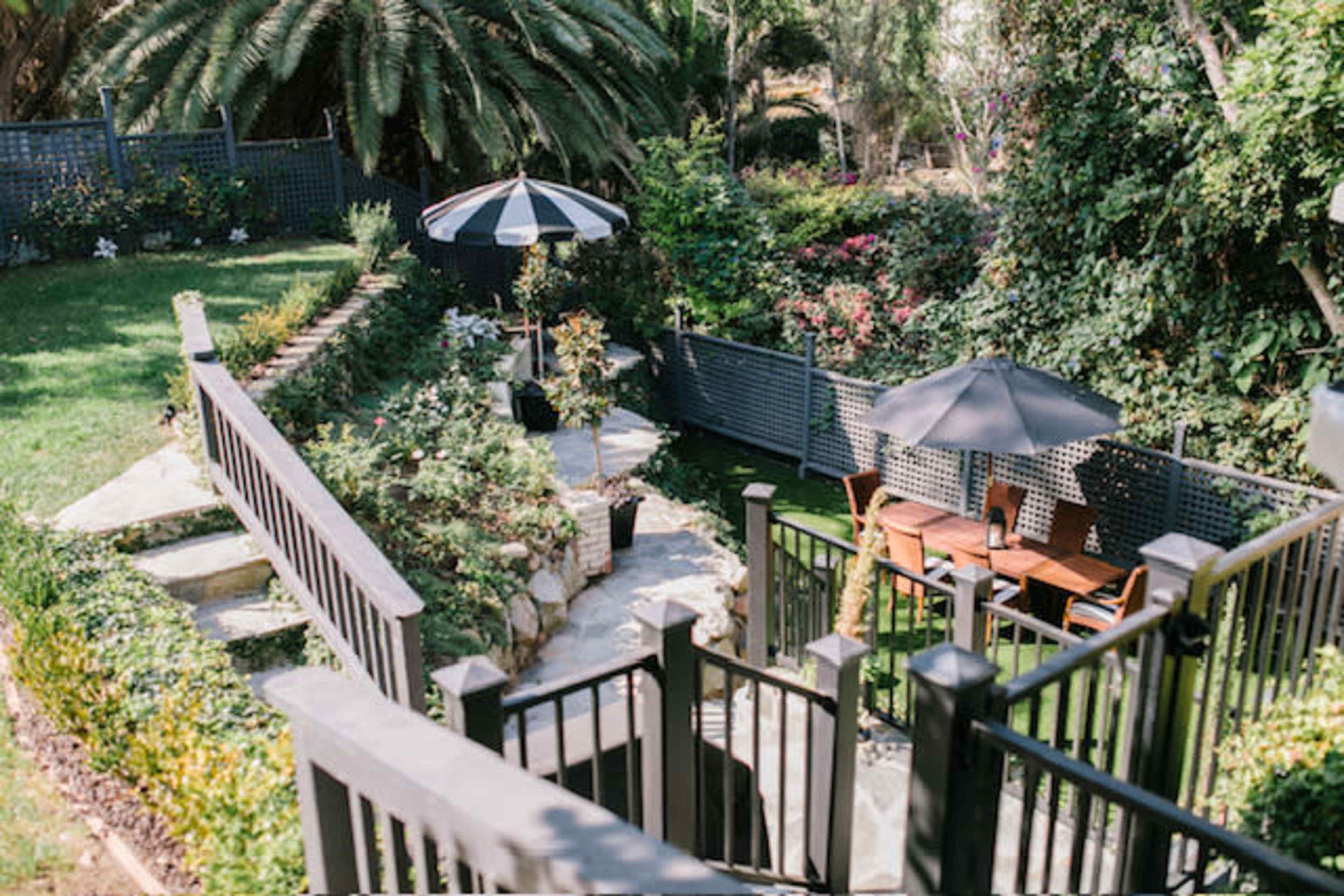 A landscaped backyard featuring a stone pathway, a dining area under black umbrellas, and lush greenery surrounding the space.
