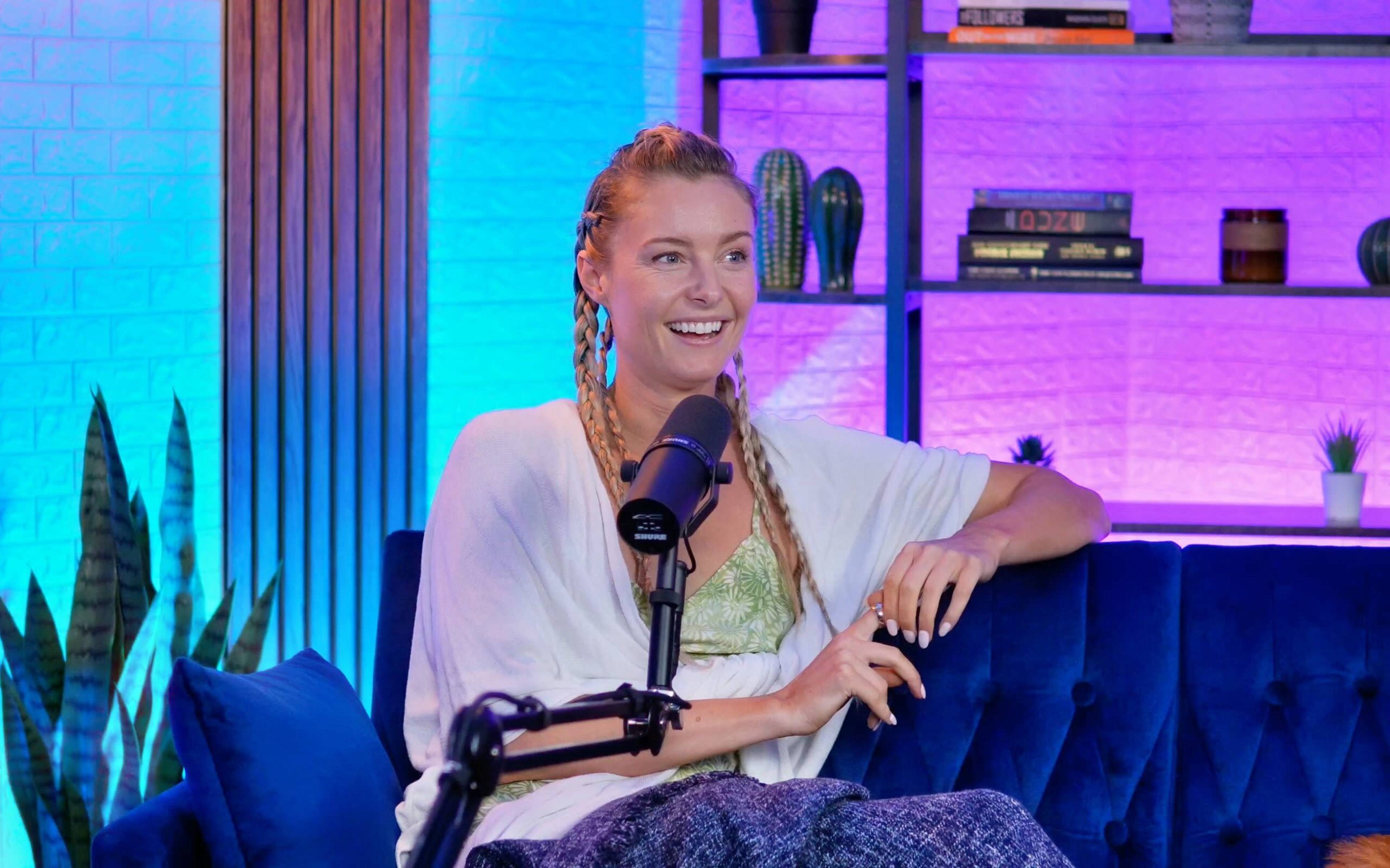 A woman with braided hair is sitting on a blue couch, smiling while speaking into a microphone in a brightly lit studio with colorful background lighting and bookshelves.