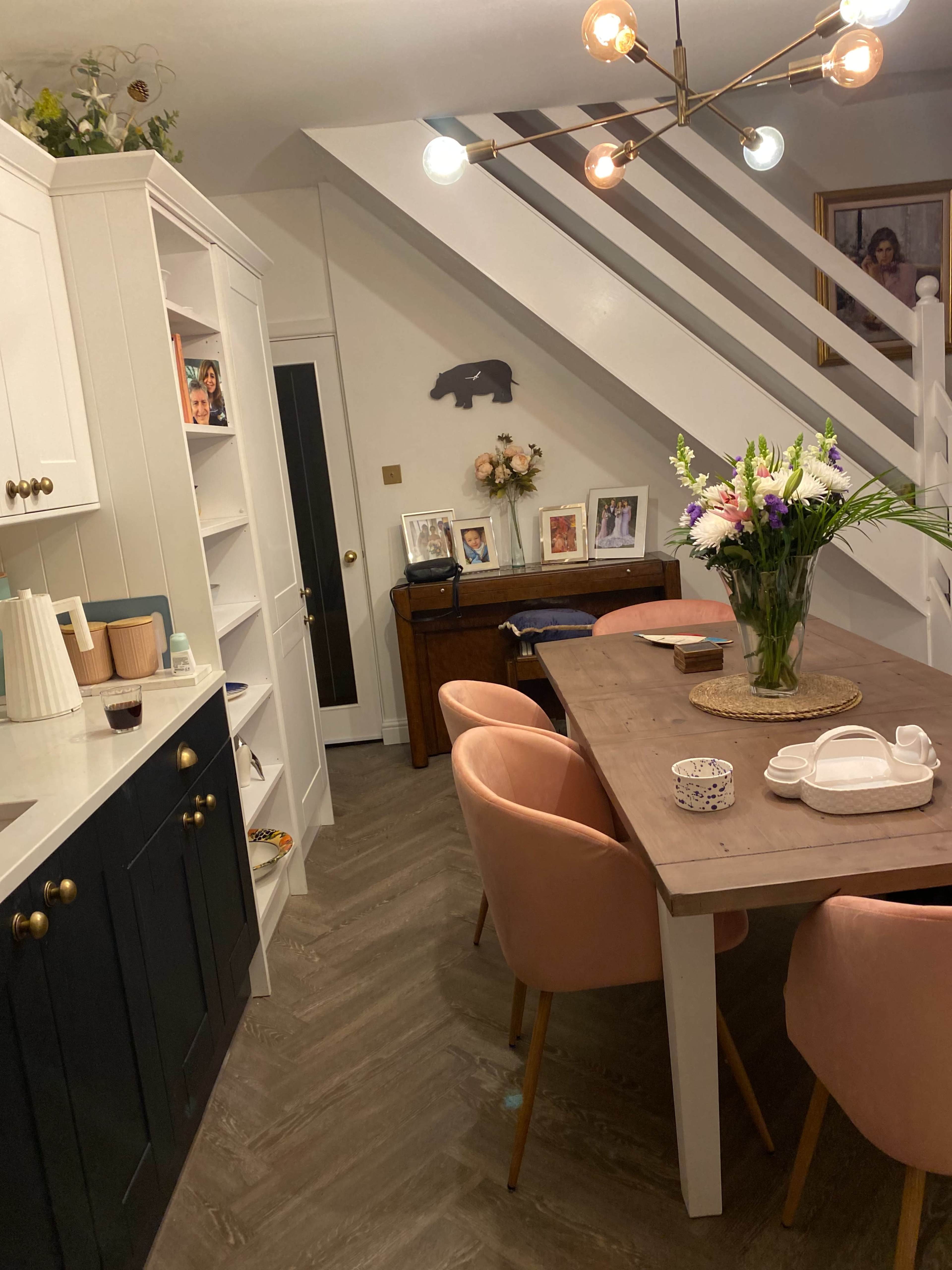 A modern kitchen-dining area featuring a wooden table surrounded by pink chairs, with a staircase and artwork in the background.