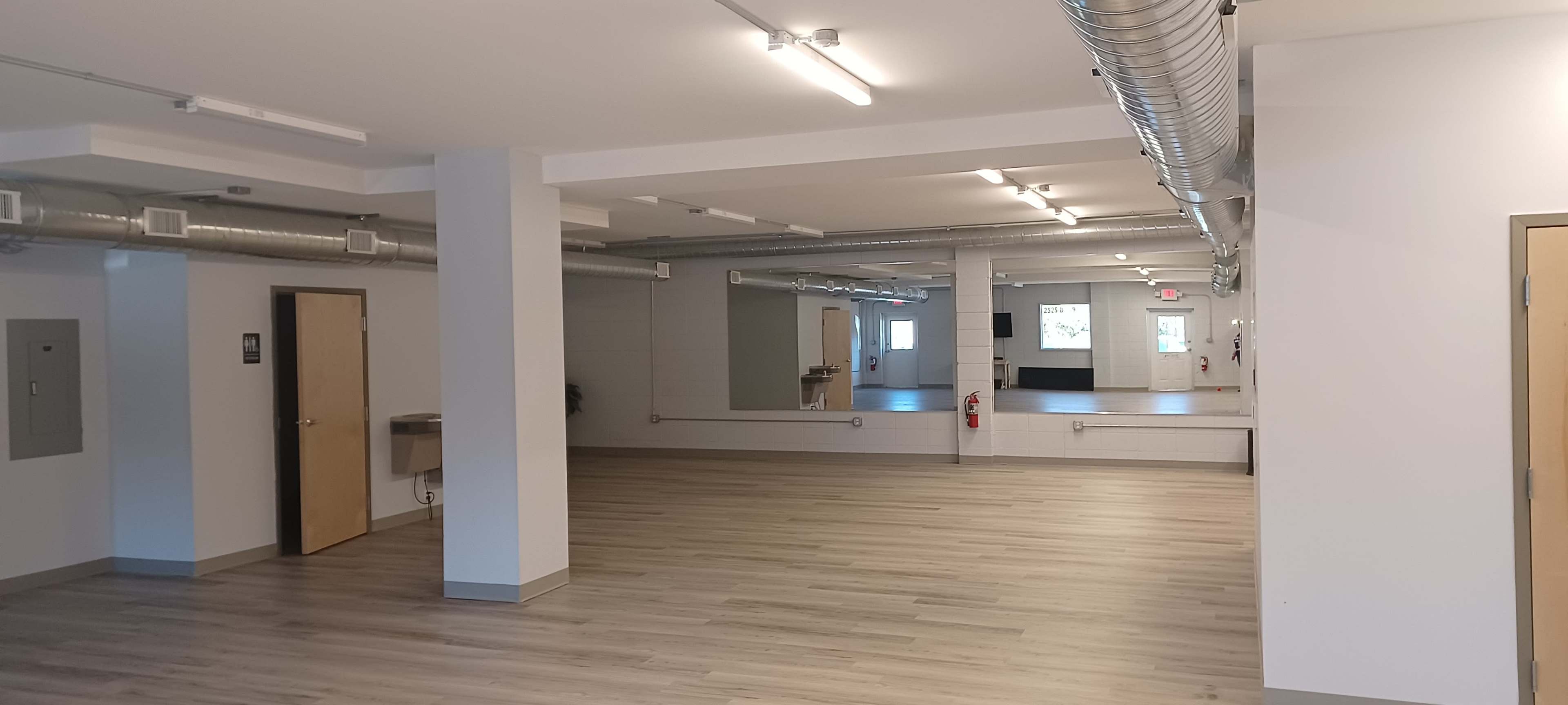 The image shows a spacious, empty room with wooden flooring, industrial lighting, and a large mirror on one wall.