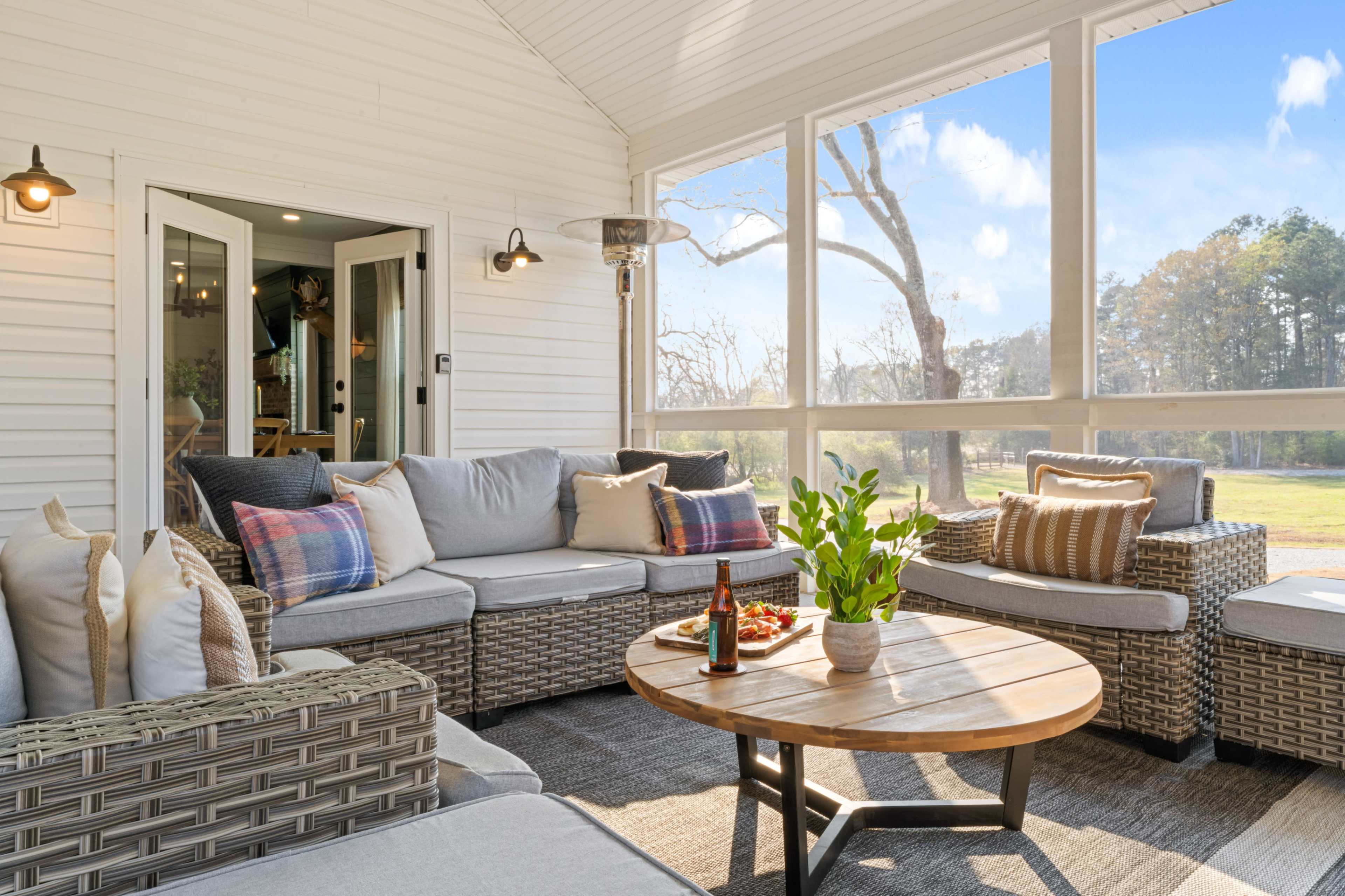 The image shows a cozy screened-in porch with gray wicker seating, a round wooden coffee table, and a view of a green landscape through large windows.