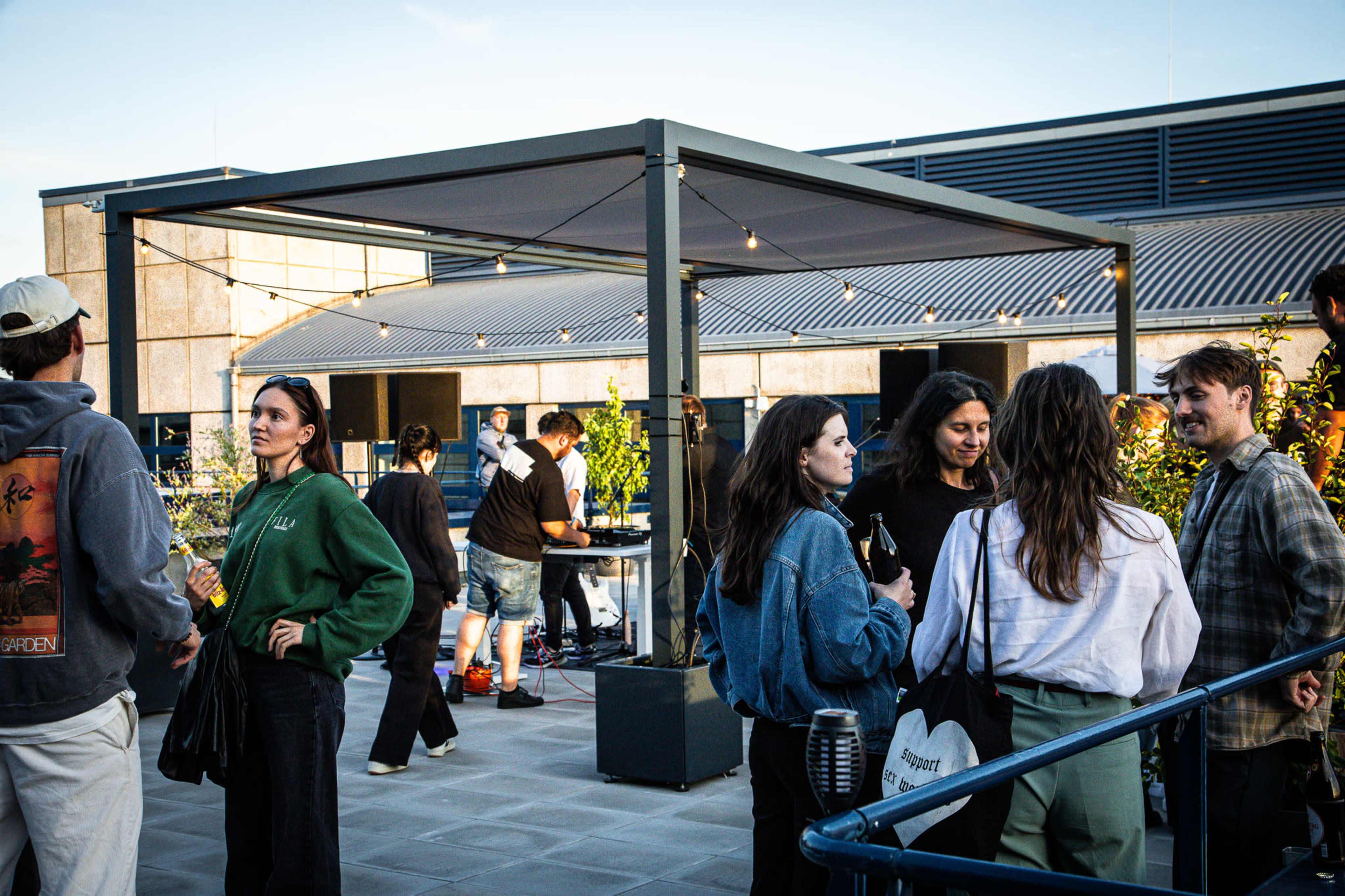 A group of people socializes on a rooftop, with some gathered under a structure while others stand around enjoying drinks.