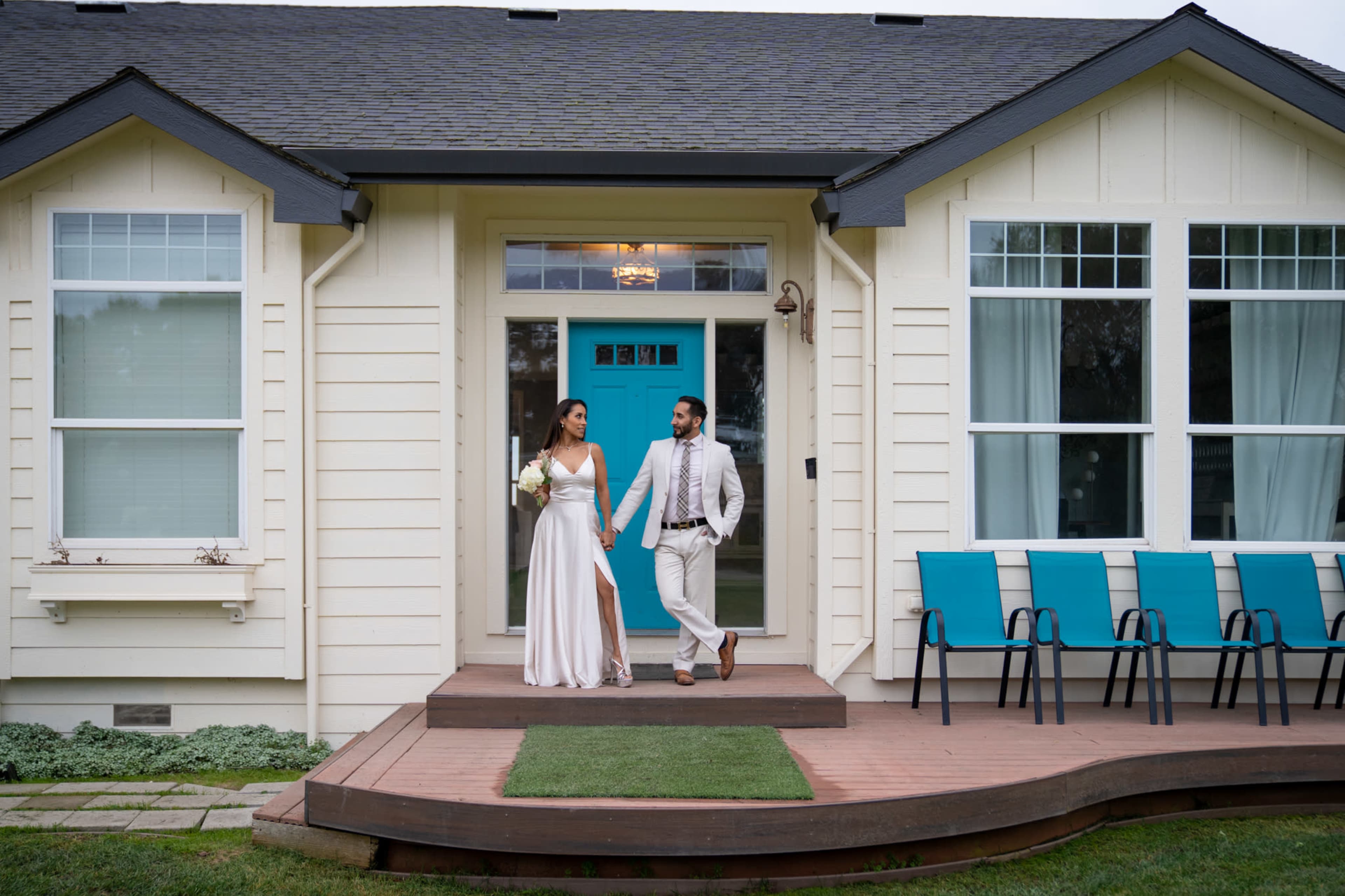 A couple dressed in formal attire is walking hand in hand on the porch of a house with teal accents and a row of blue chairs.