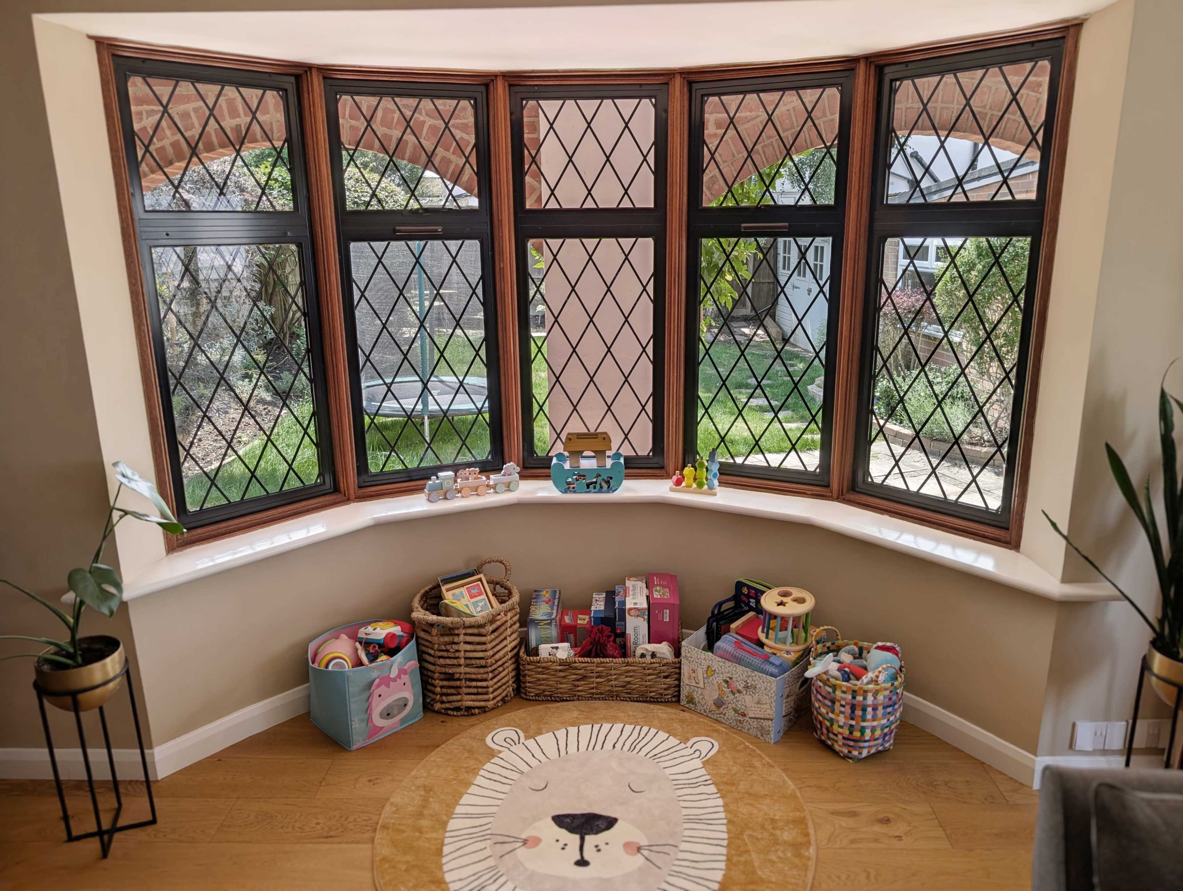 A cozy corner with large diamond-pane windows overlooks a garden, featuring baskets filled with toys and a circular rug with a lion design on the floor.